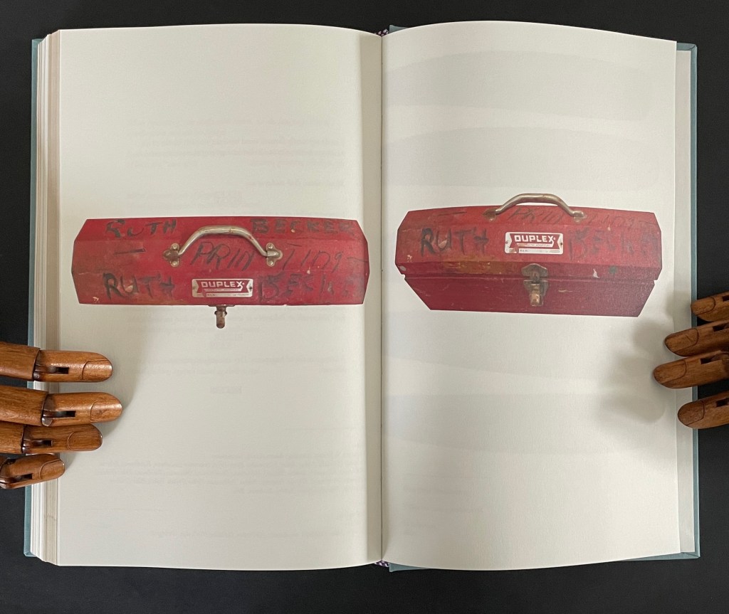 A red toolbox with the name 'Ruth' and other markings, placed open on a book's pages with wooden hands visibly holding the book.
