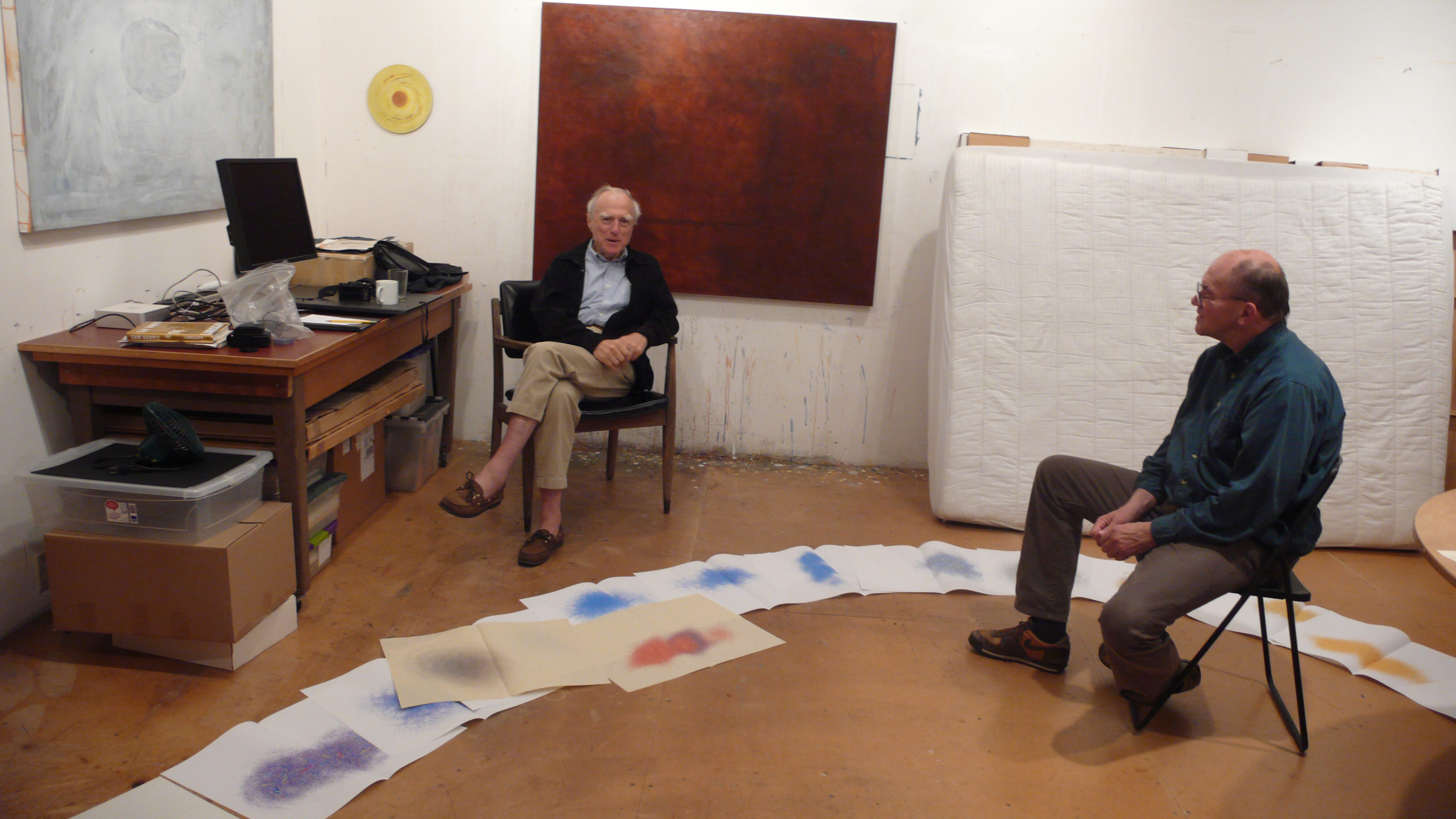 Two men seated in an art studio, discussing a series of colorful paper artworks laid out on the floor. One man sits on a chair while the other is seated on a folding chair. Behind them are boxes and a bed mattress, along with a desk containing art supplies.