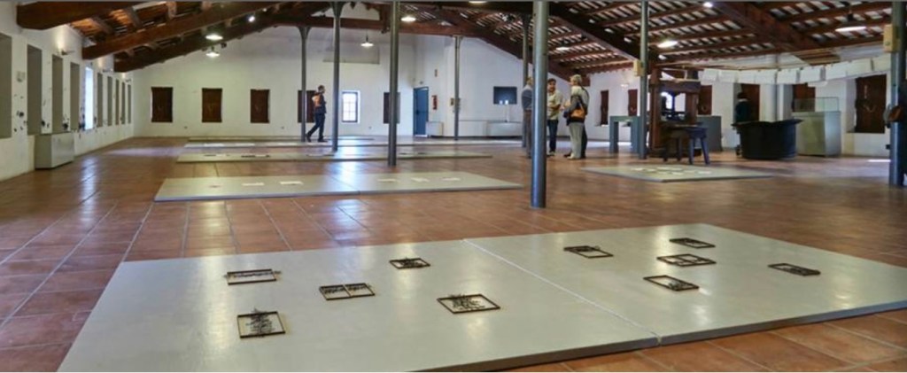 Interior view of an exhibition space with tile flooring, featuring several display panels arranged on the ground and visitors interacting in the background.