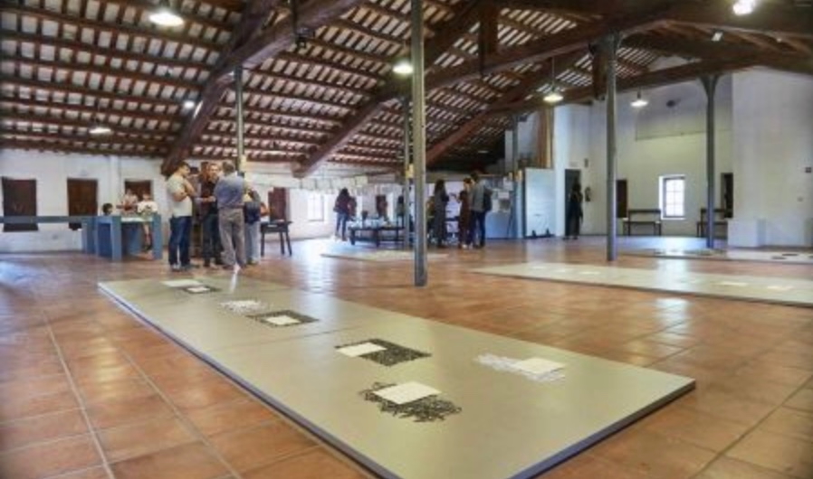 An art exhibition space featuring several display tables with artworks on a tiled floor, and visitors engaging with the exhibits in a spacious, well-lit room with wooden beams.