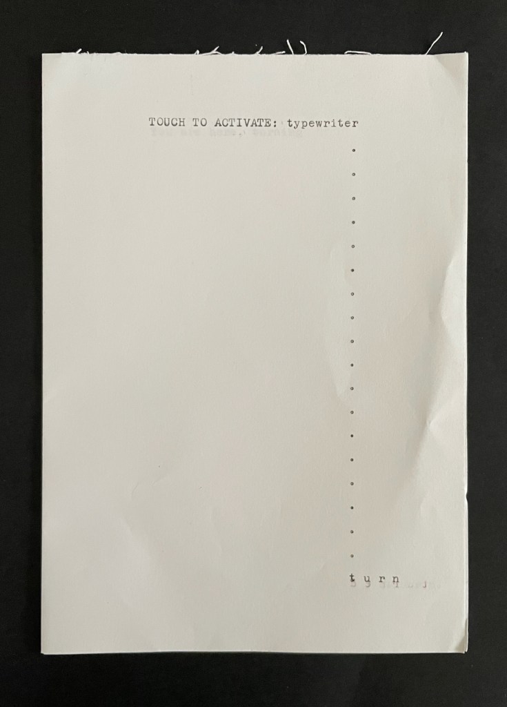 A white sheet of paper with the text 'TOUCH TO ACTIVATE: typewriter' printed at the top and a series of dots along the right side. The lower right corner has partially typed text that appears to read 'tyrn...'.