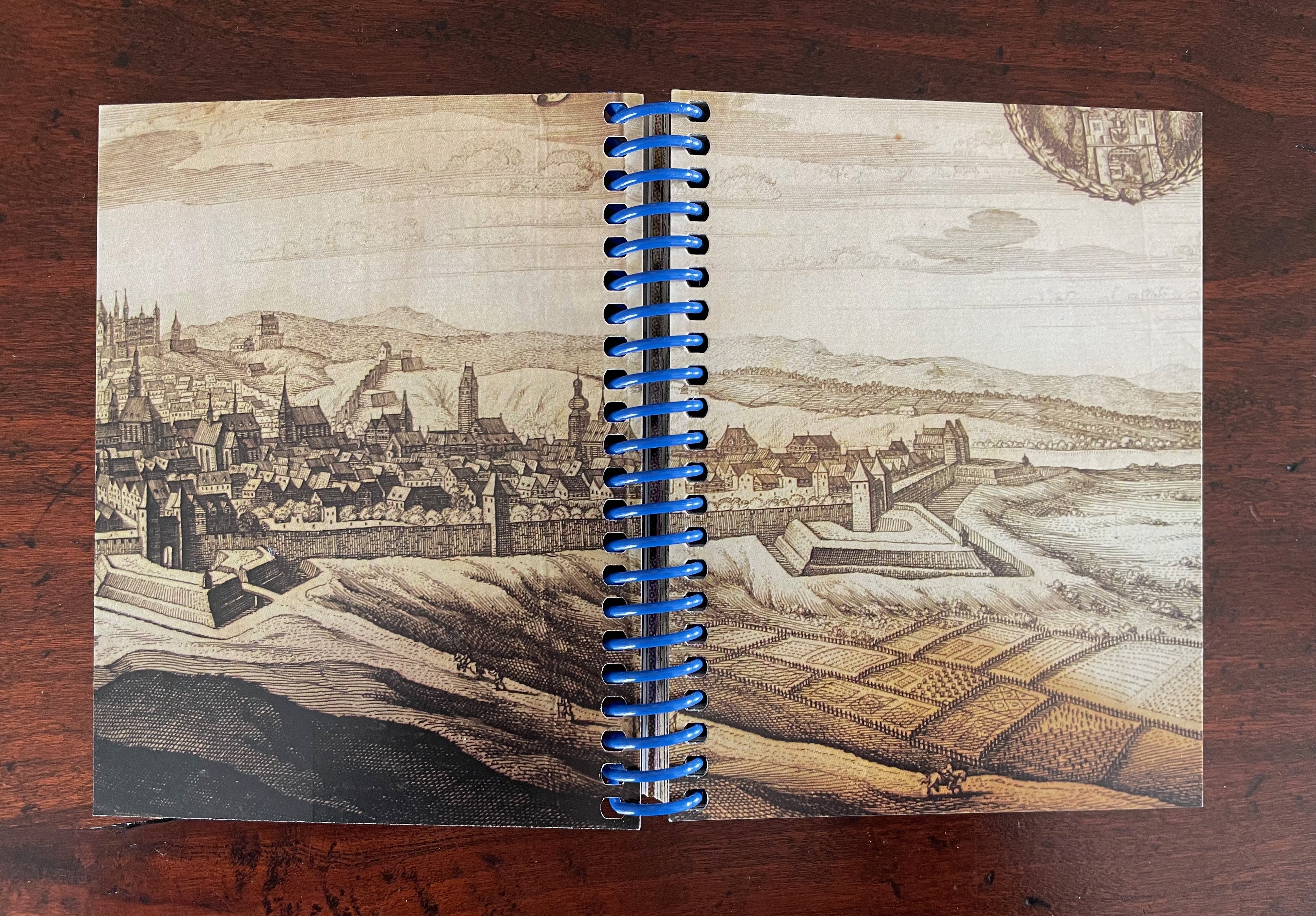 An open notebook featuring an illustrated map of a city landscape with buildings, hills, and fields, showcasing historical architecture.