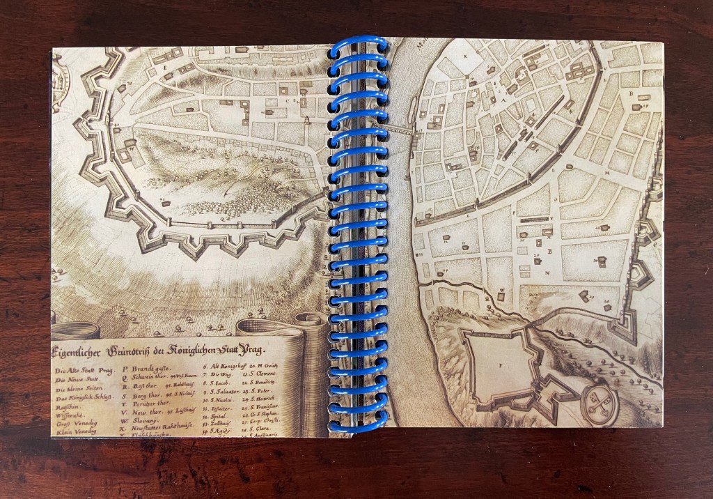 An open spiral-bound notebook displaying a historical map of a city, featuring fortifications and labeled structures. The map is in sepia tones, with a list of locations and names at the bottom.