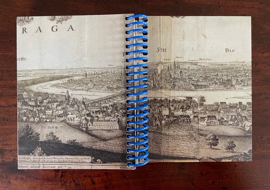 An open notebook with a spiral binding, displaying an old illustrated map of a city, featuring buildings and a river.