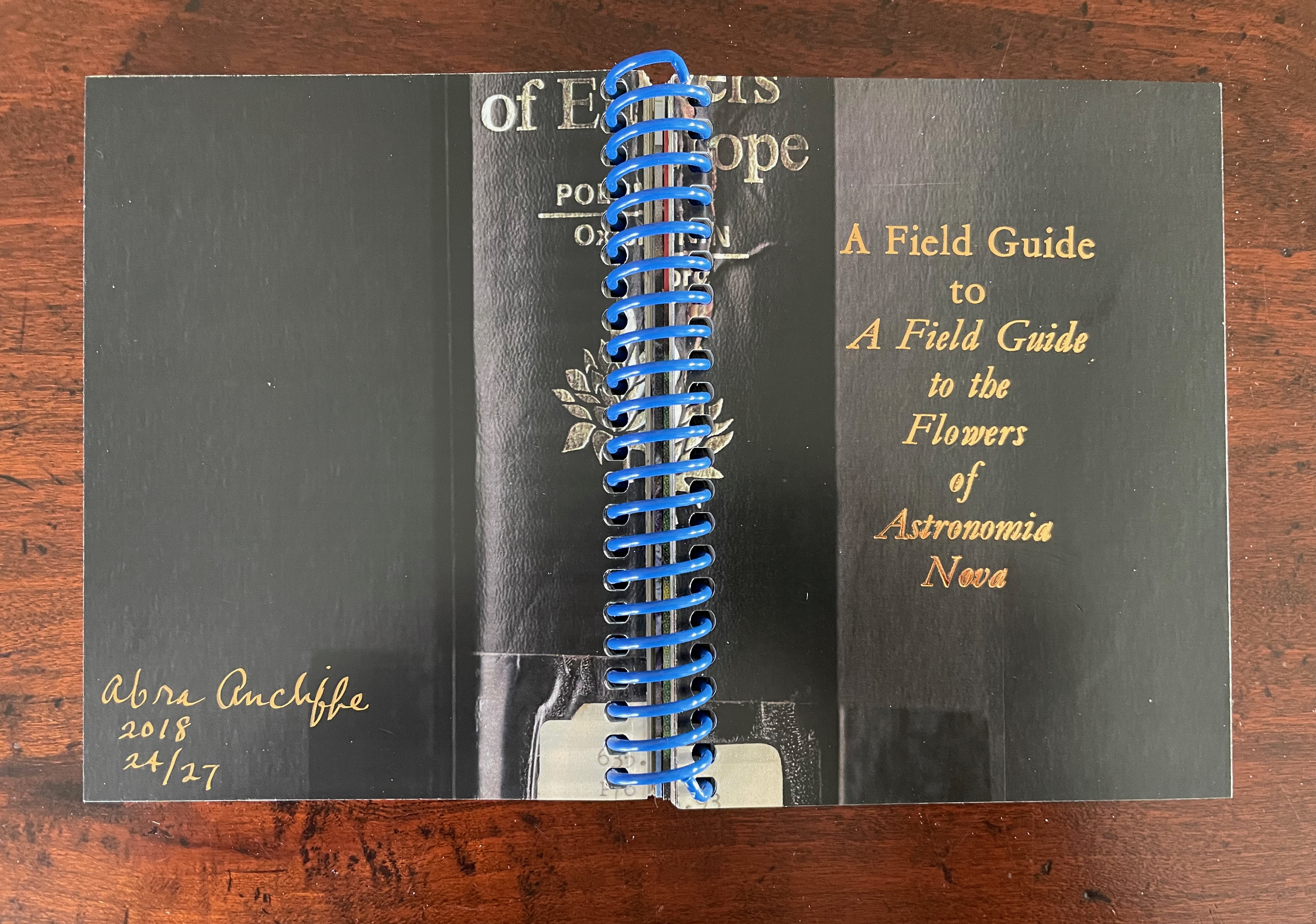 A close-up of an open field guide book titled 'A Field Guide to the Flowers of Astronomia Nova' with blue spiral binding and a signature by the author, Abra Anchifé, dated 2018.