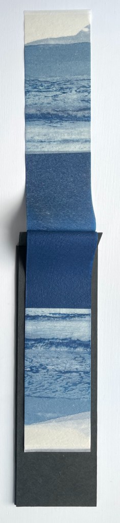 A vertical strip of paper featuring a gradient of blue tones, depicting ocean waves and shorelines. The strip is placed within a black holder.