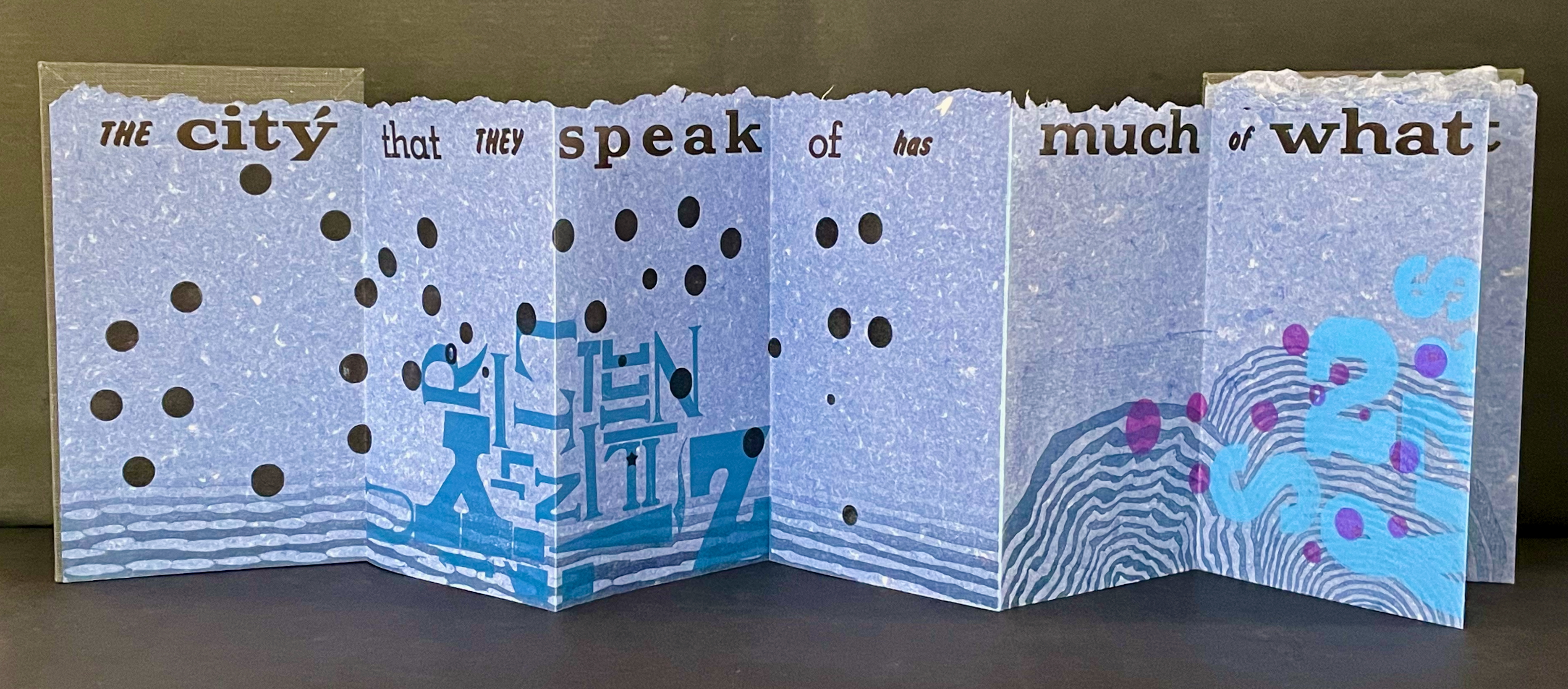 An artistic fold-out book with layered translucent pages, featuring text and abstract designs in blue, black, and purple. The text reads: 'THE city that THEY speak of has much of what.' The background is a light blue texture with circular cutouts.