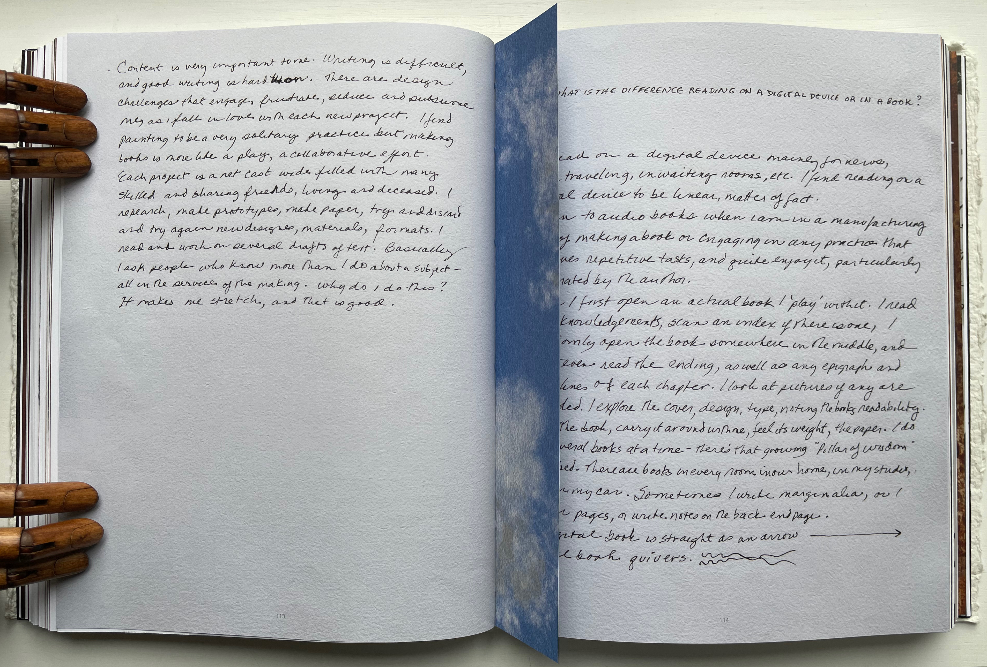 An open book displaying handwritten notes on the left and right pages, with a wooden hand sculpture holding the left page. The text discusses the differences between writing on various media and the experience of reading a physical book versus a digital device.