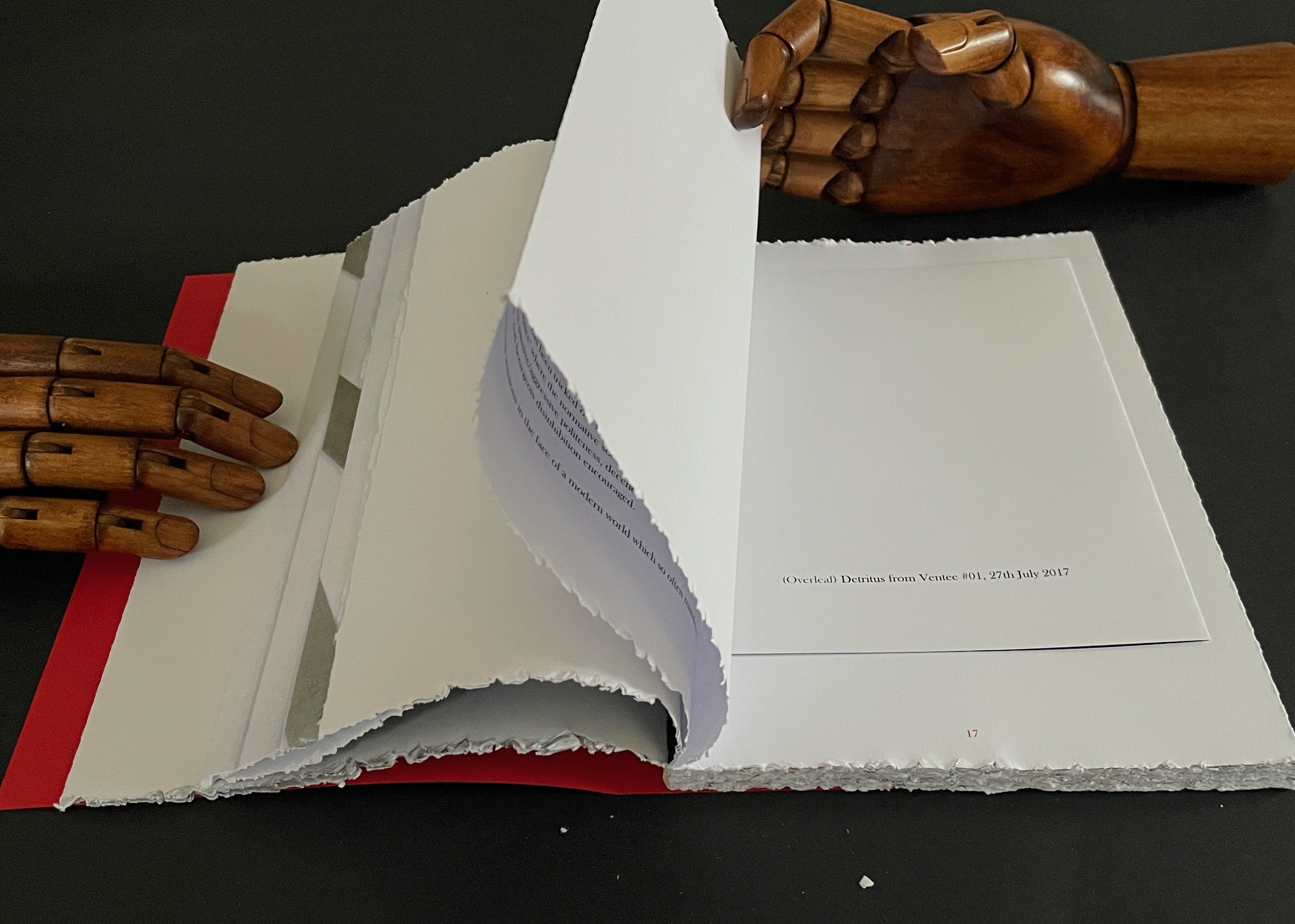 A partially opened art book resting on a dark surface, with wooden mannequin hands holding the pages. The pages feature a mix of blank sheets and text.
