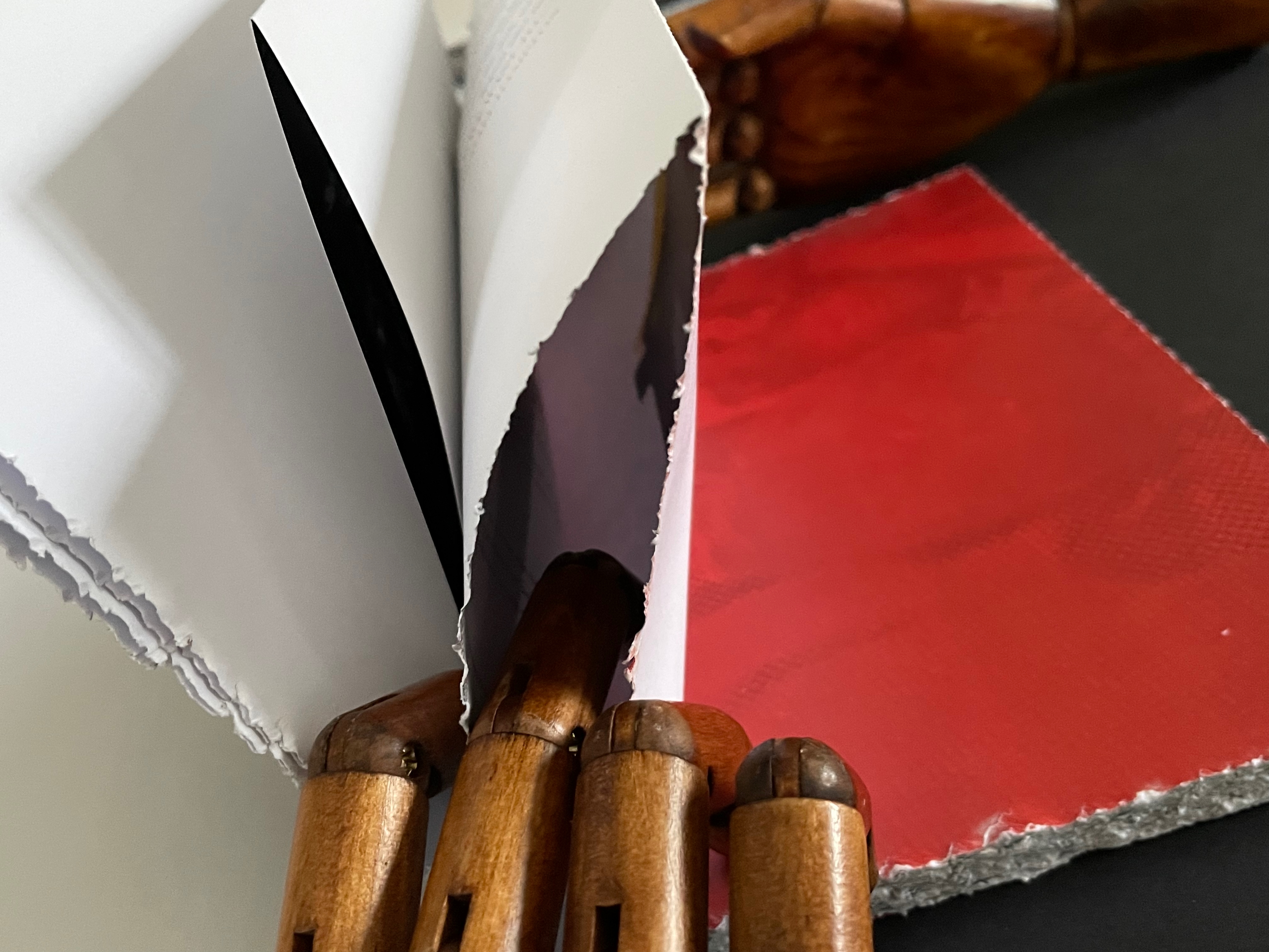 Close-up of a partially opened book with blank pages, revealing a red textured material and a wooden art mannequin hand in the background.