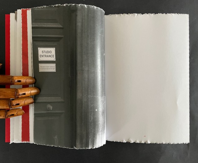 An open book displaying a black and white photo of a studio entrance door on the left page, with the text 'STUDIO ENTRANCE' on a sign. The right page is blank with torn edges. A wooden hand is partially visible on the left, holding the book.