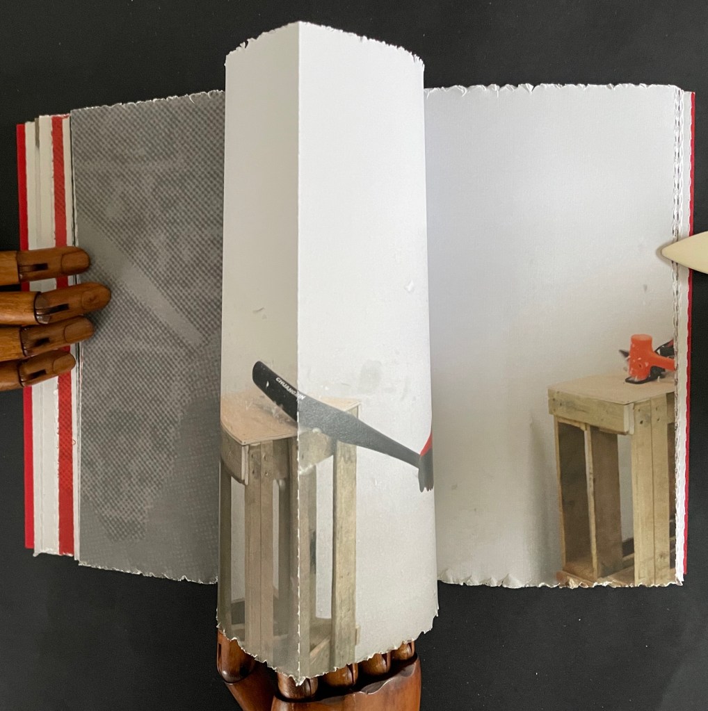 An open book displaying images of a workshop environment, featuring wooden crates and tools. A wooden hand is holding the book, showcasing the textured pages.