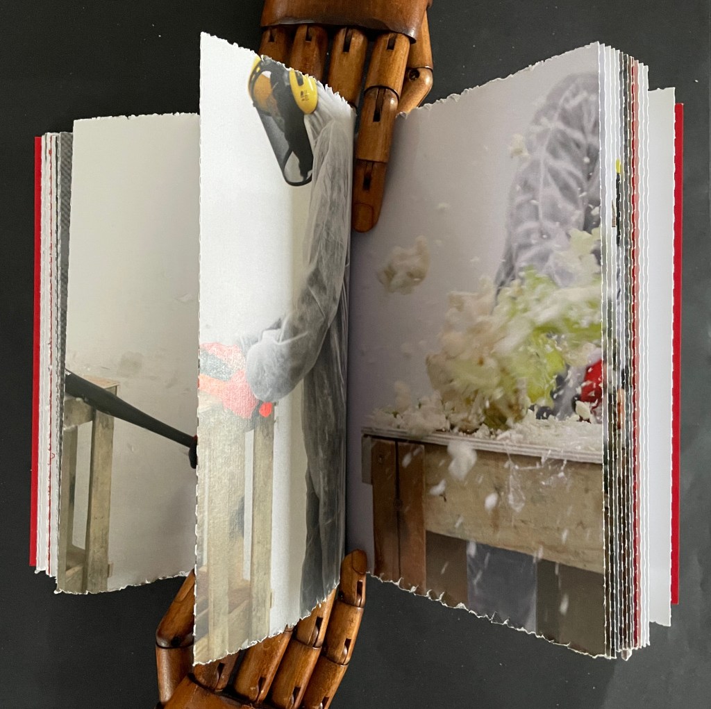 An open book displaying colorful images on the pages, being held by a wooden hand model. The left page shows a person in protective gear working with a tool, while the right page captures a dynamic scene of an object disintegrating, with bits flying out.