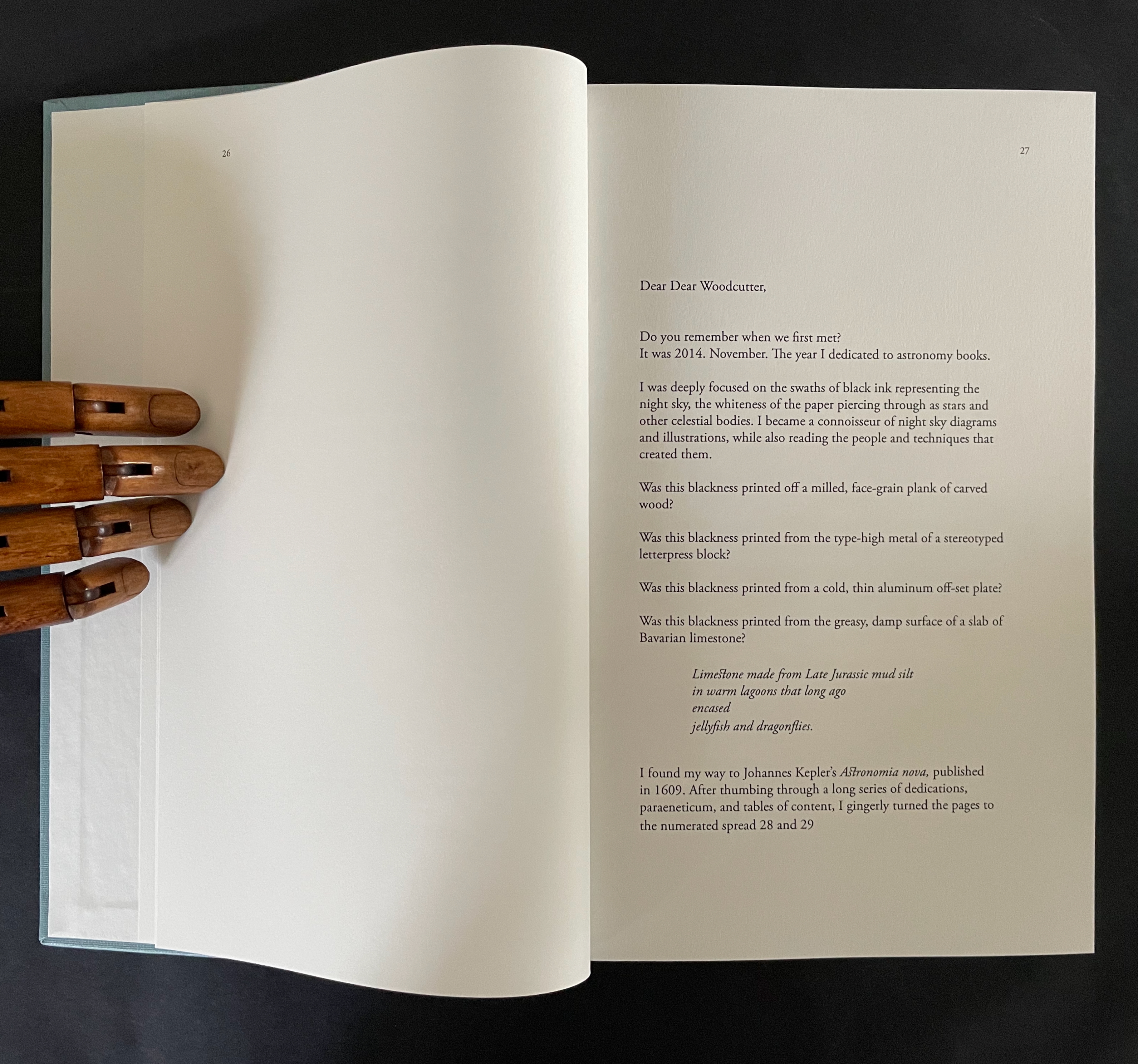 An open book showing a partially visible page with text on the right and a blank page on the left. A wooden hand puppet is holding the book. The text discusses memories related to astronomy and printing techniques.