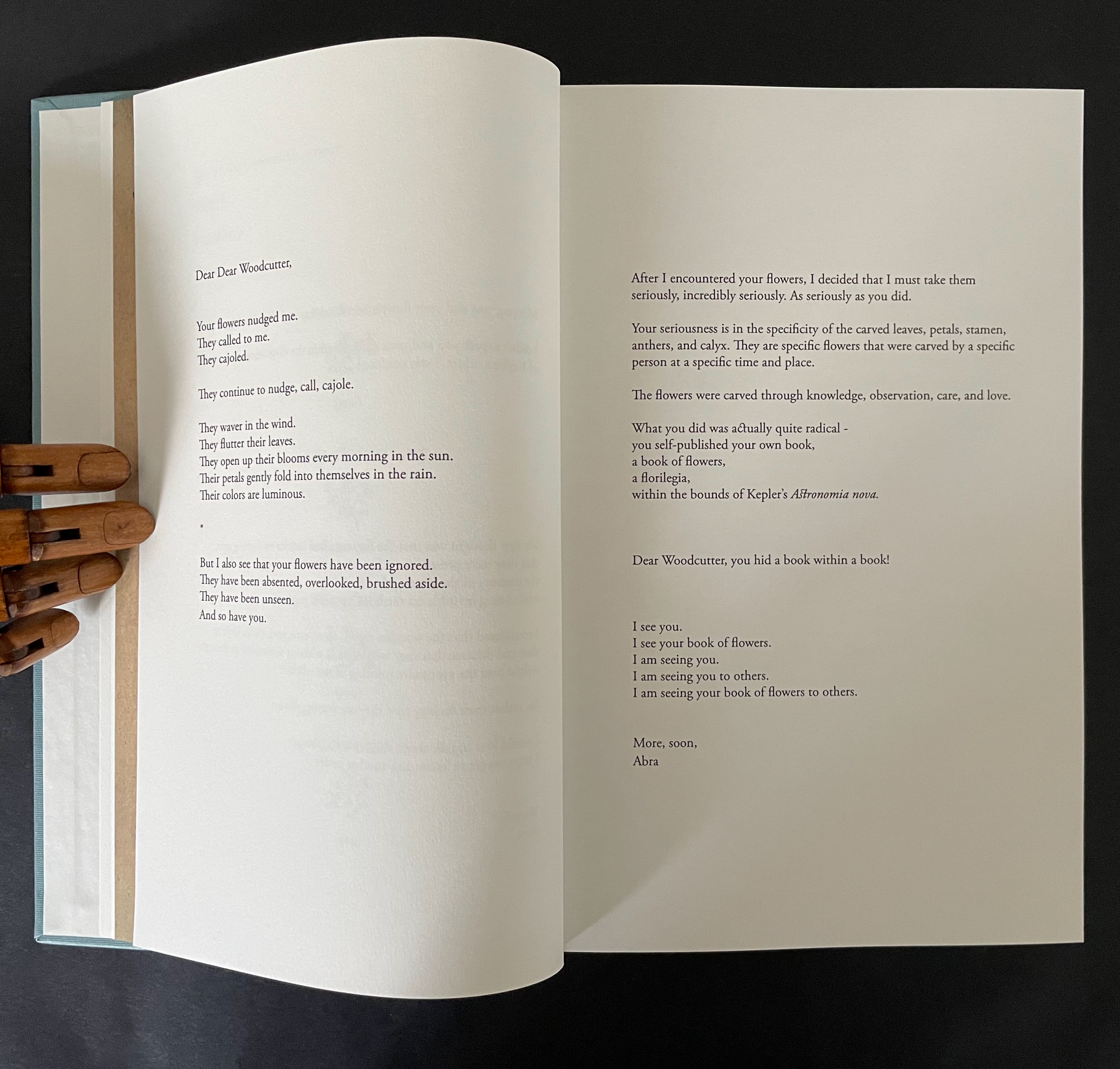 An open book displaying a handwritten letter on the left page and printed text on the right page, accompanied by a wooden hand model resting on the edge.