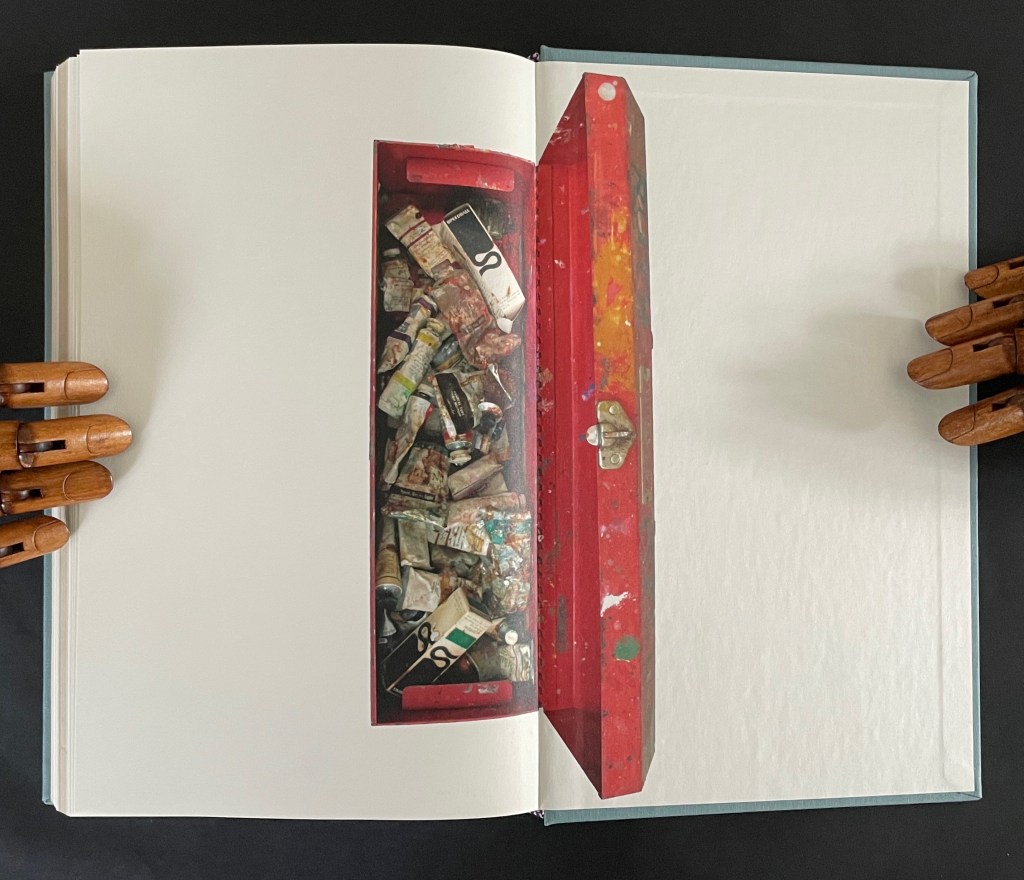 An open red toolbox filled with paint tubes on a page of a book, held by wooden hands.
