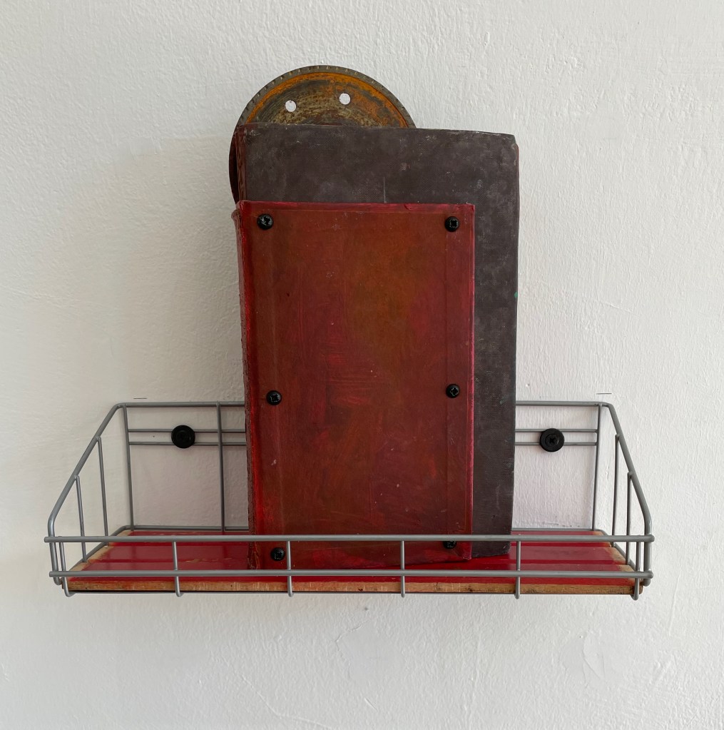 A metal wire shelf holding two stacked books, one with a red cover and the other with a dark cover, mounted against a white wall.