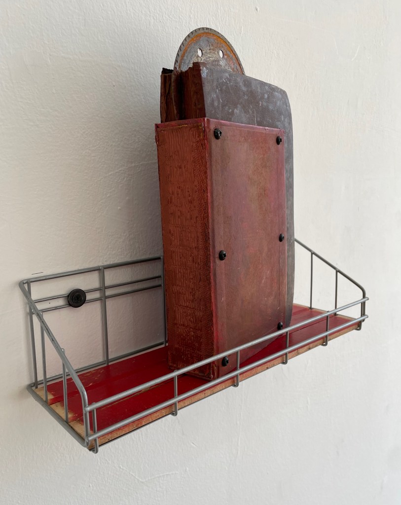 A metal wall-mounted shelf holding a rust-colored object and a wooden board, set against a light-colored wall.