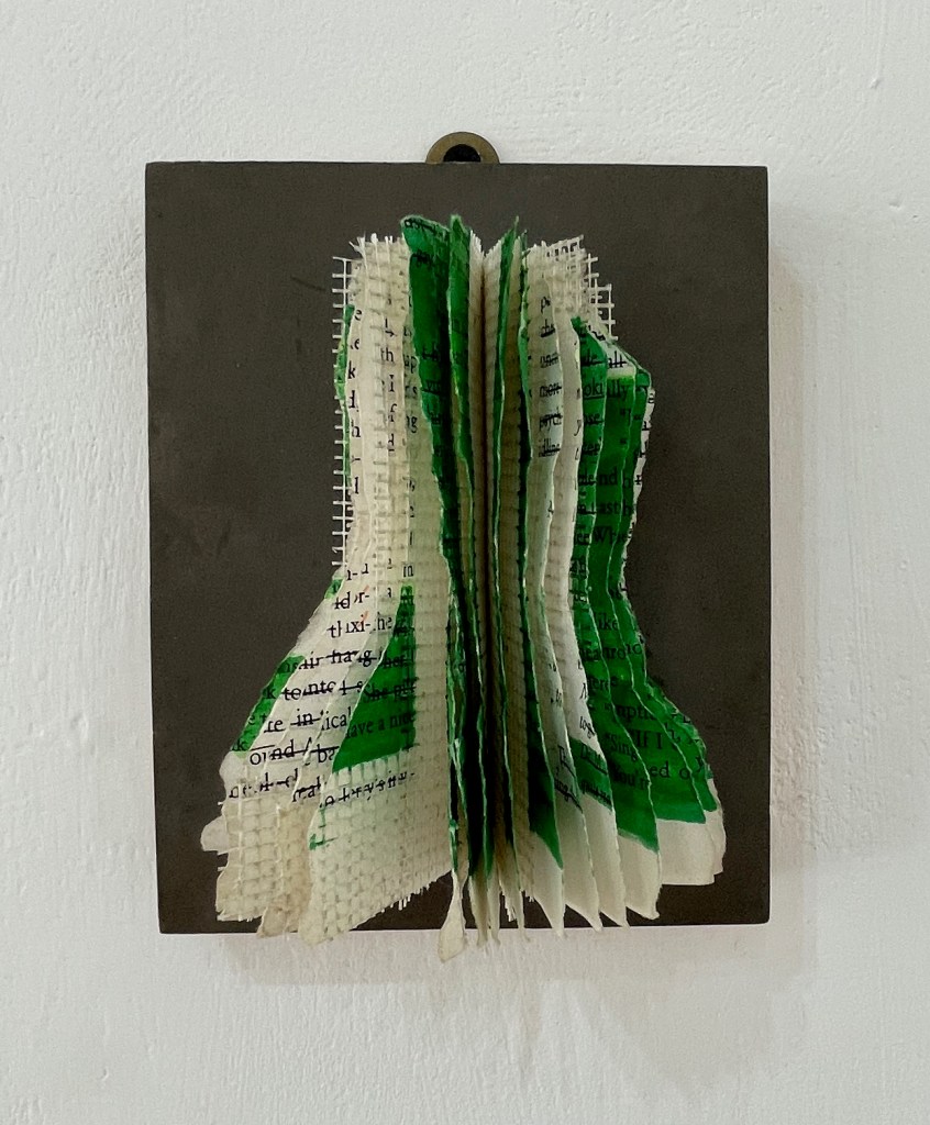 Art piece featuring layered, crinkled pages in green and white, mounted on a grey background.