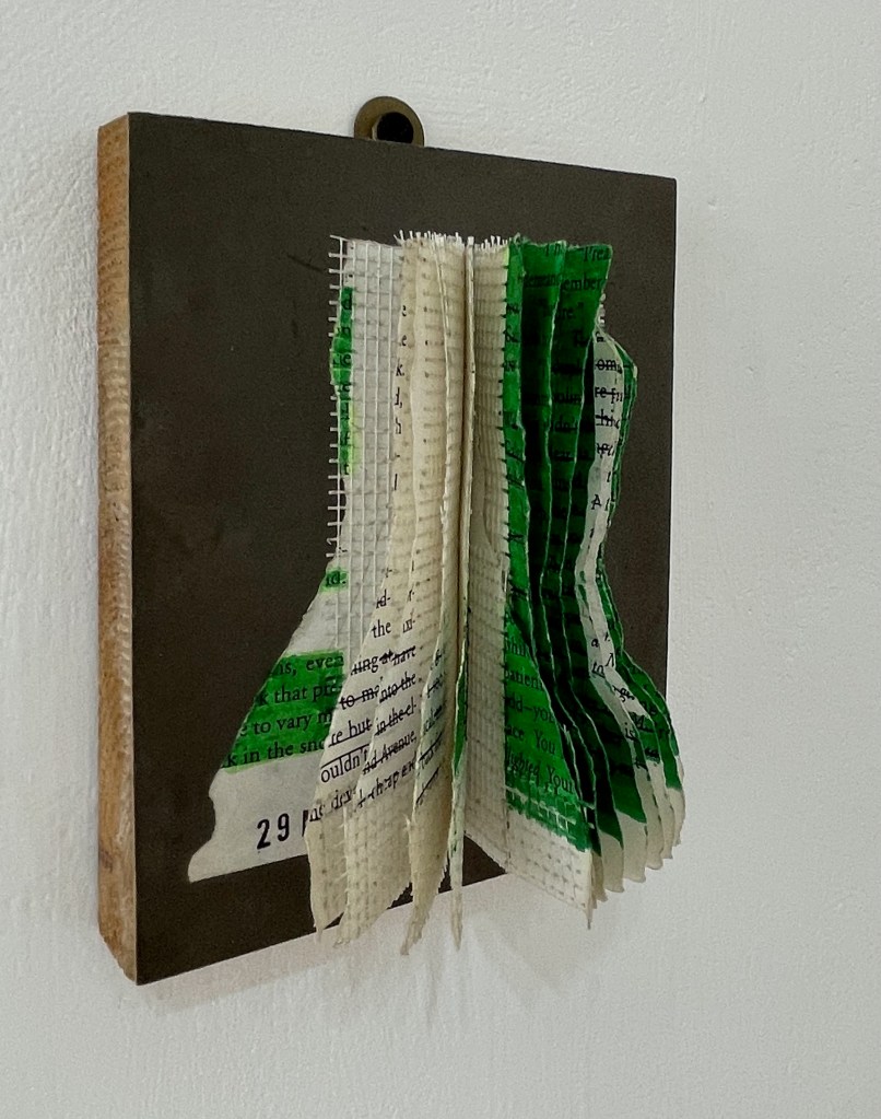 An artistic installation featuring layered pieces of paper in green and white, attached to a dark background board. The paper has text printed on it and appears to be creatively manipulated, creating a 3D effect.