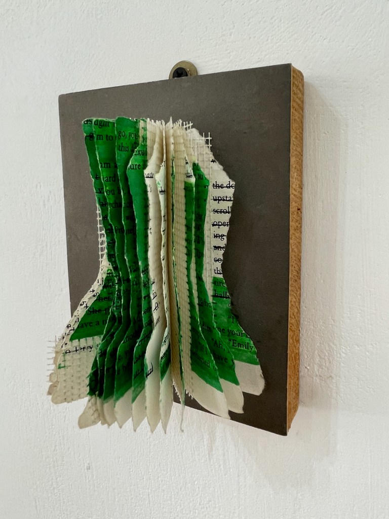 An artistic installation featuring layered pages from a book, with green highlights and a textured surface, mounted on a wooden base against a white wall.