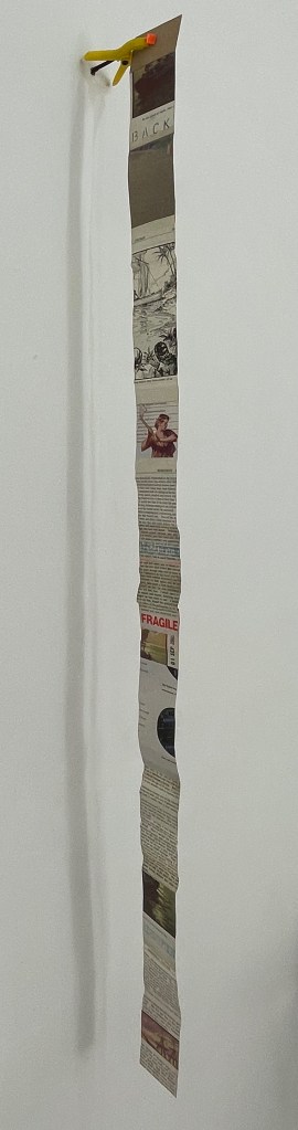 A vertical strip of paper attached to a wall, featuring various printed images and text, including illustrations and labels, with a small yellow clip at the top.