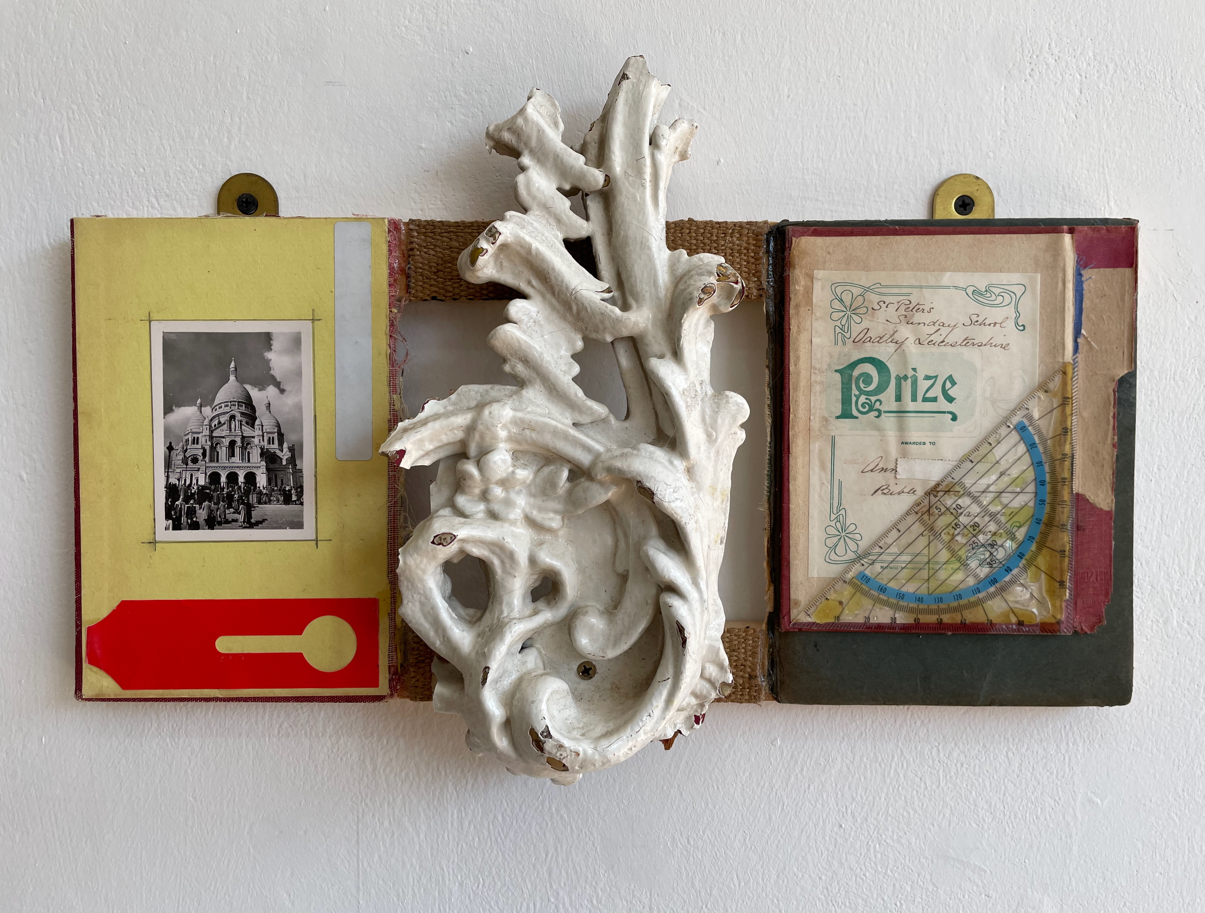 A wall-mounted display featuring a white ornamental piece alongside a black and white photo of a building, a yellow page with a red tag, and a prize certificate with a protractor.