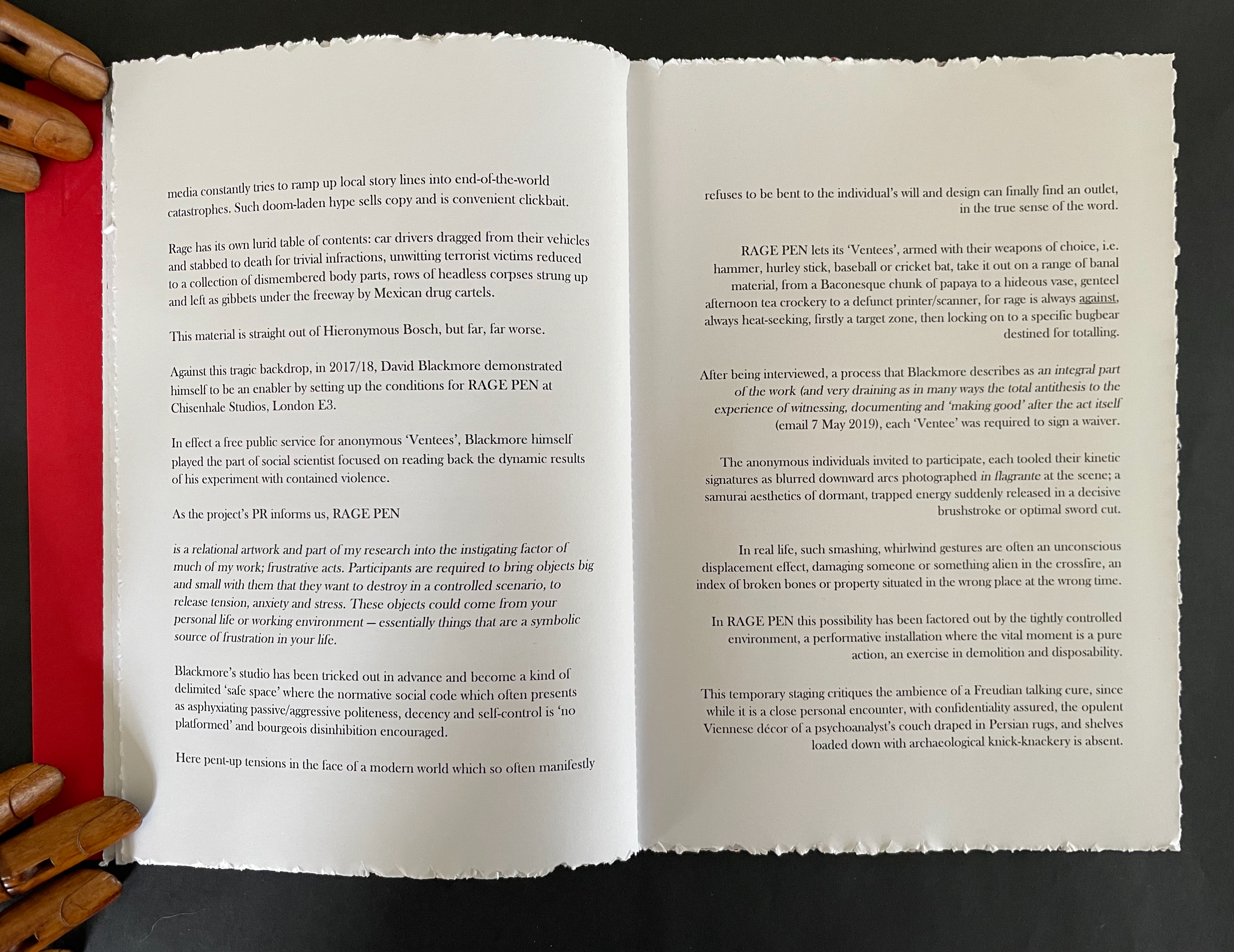 An open book with torn edges on a black surface, one page displaying printed text and the other side showing a red background. A wooden hand is partially visible on the left, suggesting a creative or artistic theme.