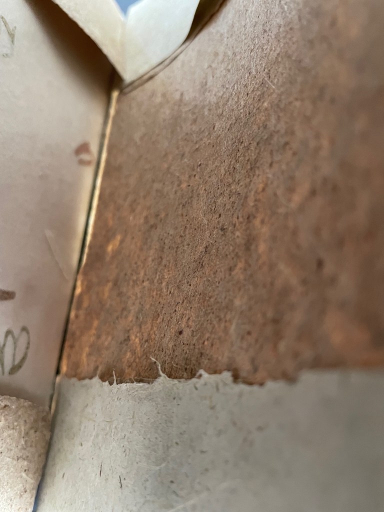 Close-up view of textured brown material with a torn edge, partially revealing a light-colored paper underneath.