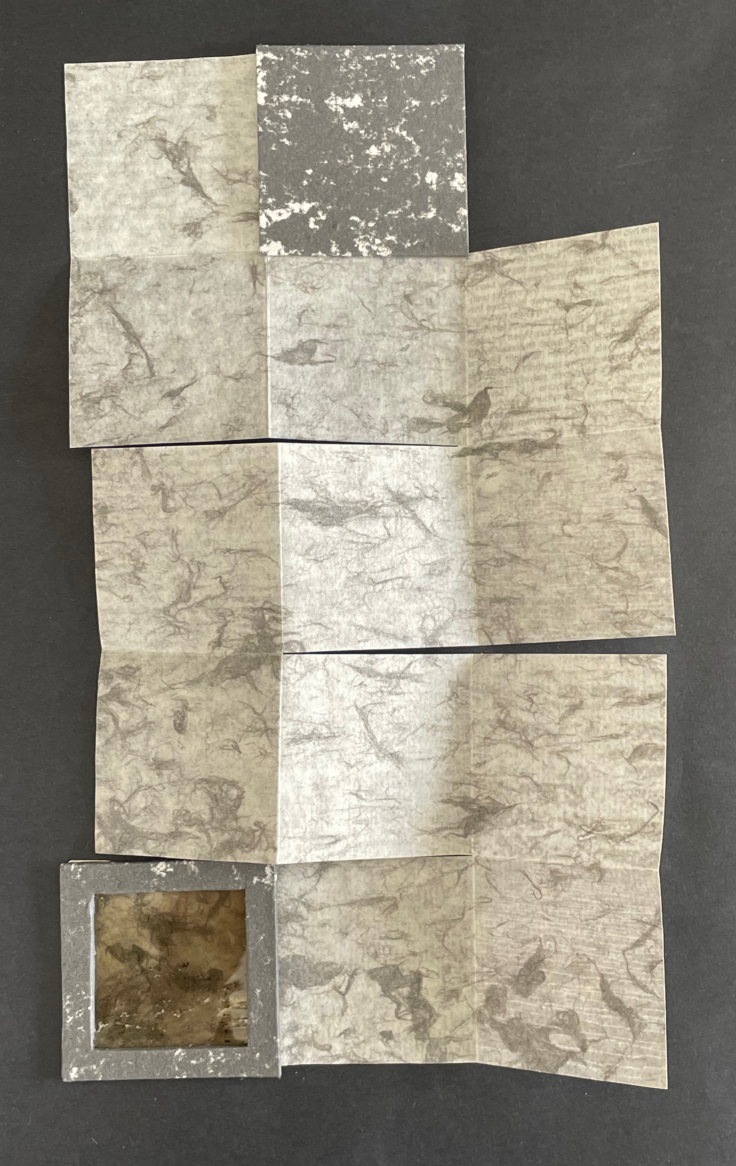 A layered composition of various textured papers in shades of gray and beige, arranged in a unique pattern, with a small square frame integrated into the design.