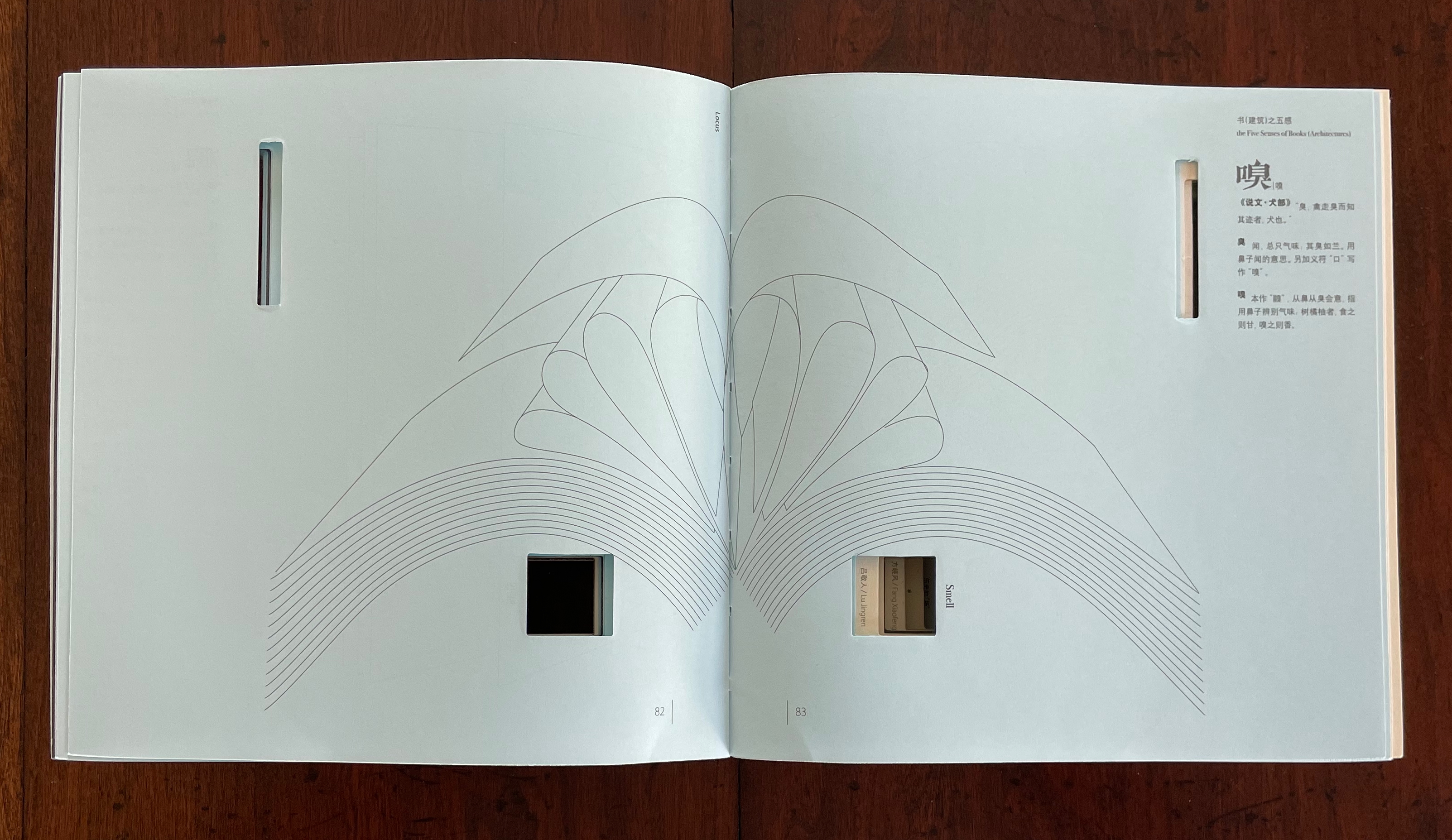 An open book displayed on a wooden surface, featuring intricate line drawings and cutouts on its pages. The left page shows curved shapes and lines, while the right page contains text in a different language along with a visible cutout.