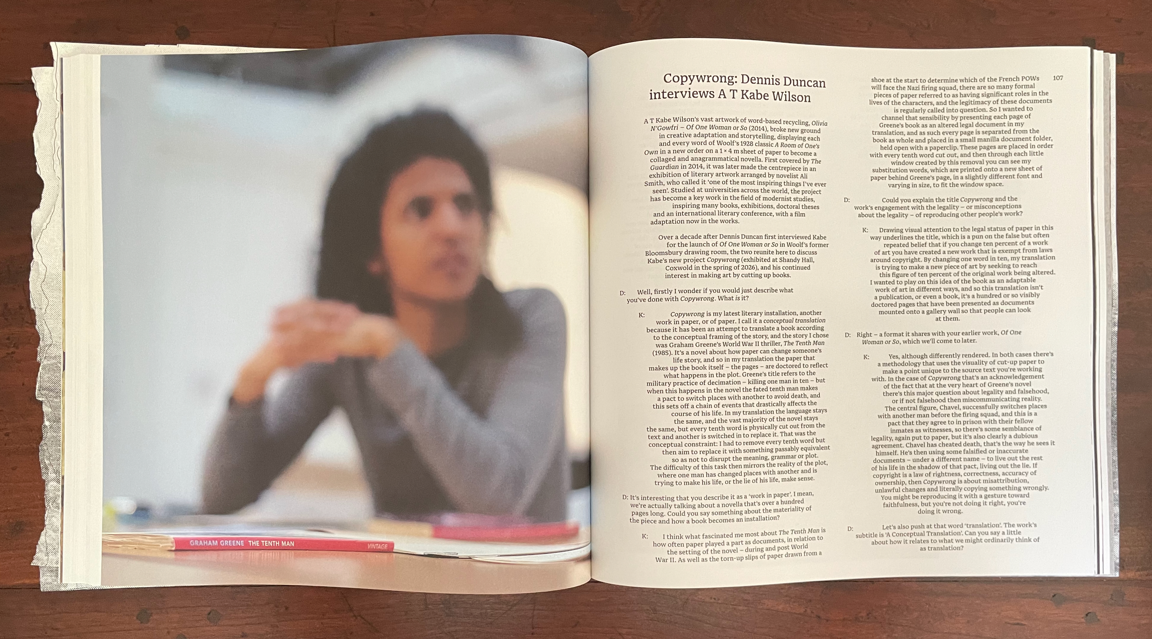 A close-up view of an open book featuring an interview between Dennis Duncan and T Kabe Wilson on the left page, with blurred focus on a person who appears to be deep in thought. The right page contains text discussing concepts of copyright and interpretation in literature. A partially visible book titled 'The Tenth Man' by Graham Greene is seen in the foreground.