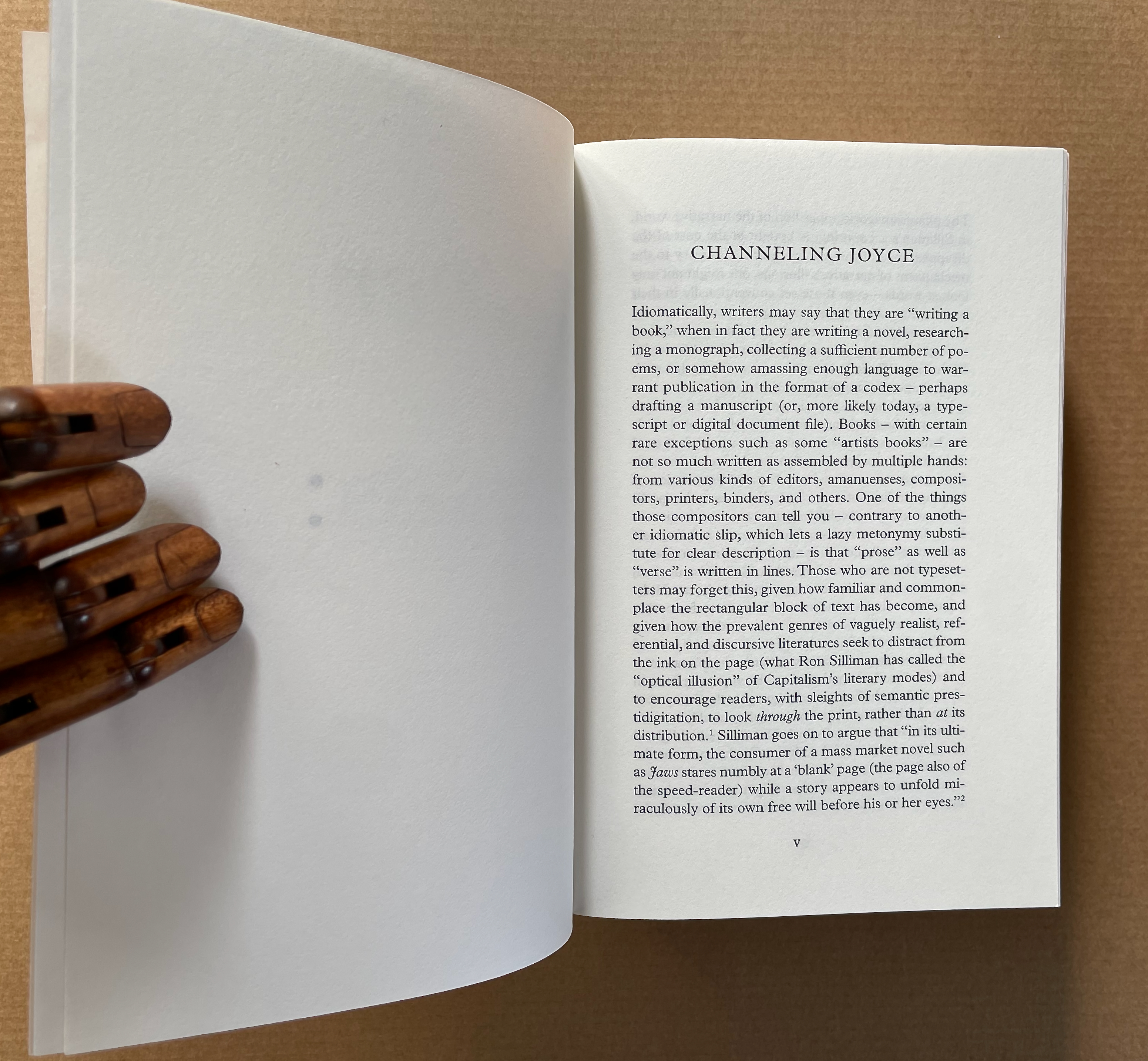 An open book displaying text on a page titled 'CHANNELING JOYCE,' with a wooden articulated hand resting on the left side.