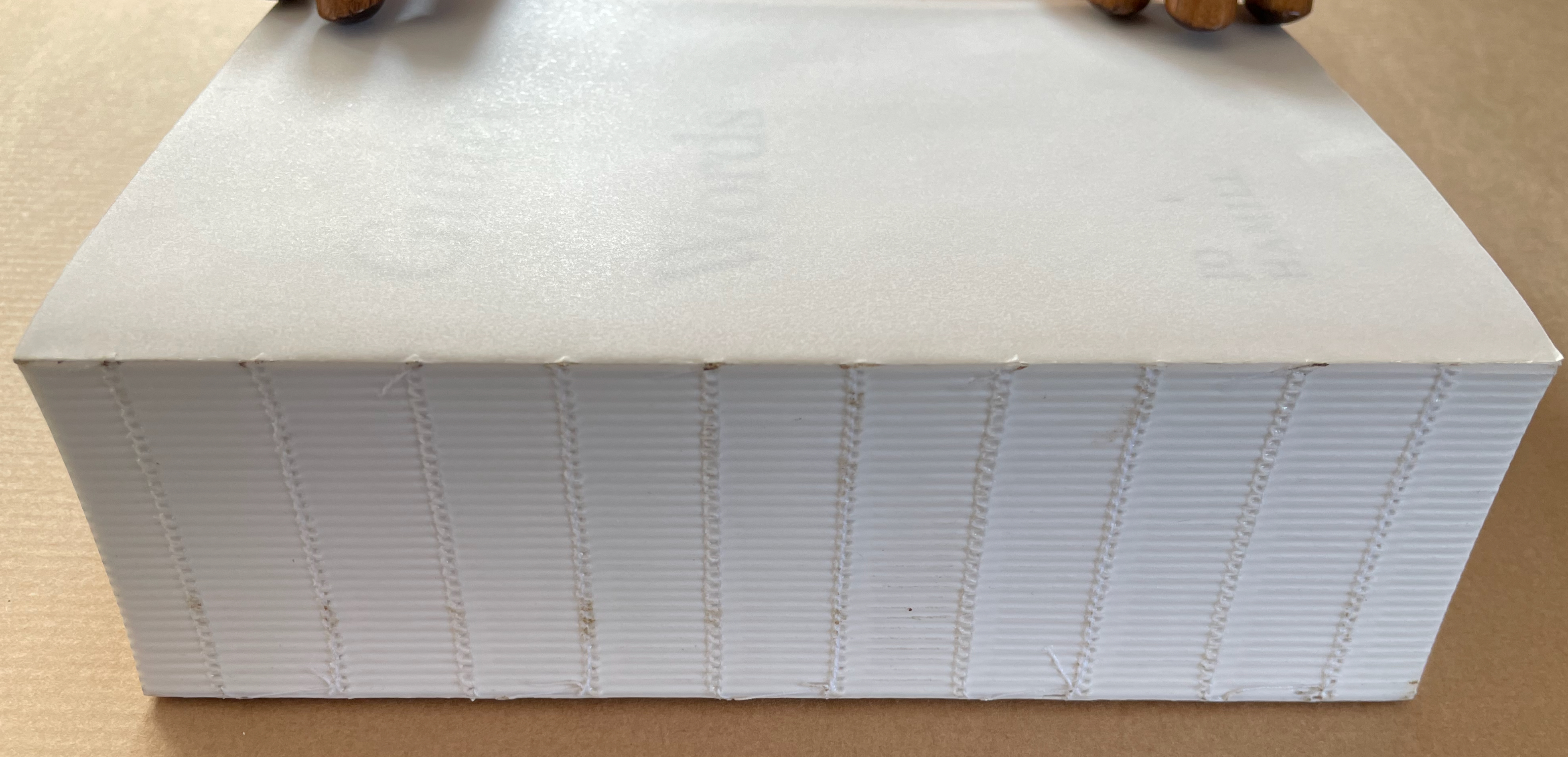 A close-up view of the edge of a thick, white book with a textured spine. The book is partially open, resting on a brown surface, with two wooden hands on top.