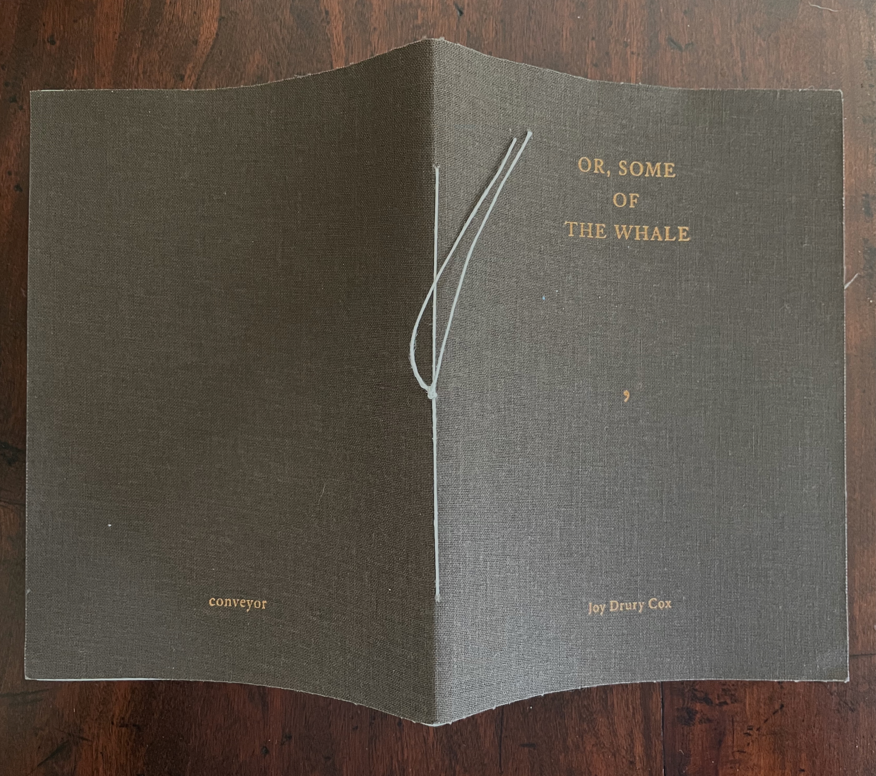 A closed book with a brown cover featuring the title 'OR, SOME OF THE WHALE' and the author's name 'Joy Drury Cox' embossed in gold lettering. The book is tied with a light blue string.