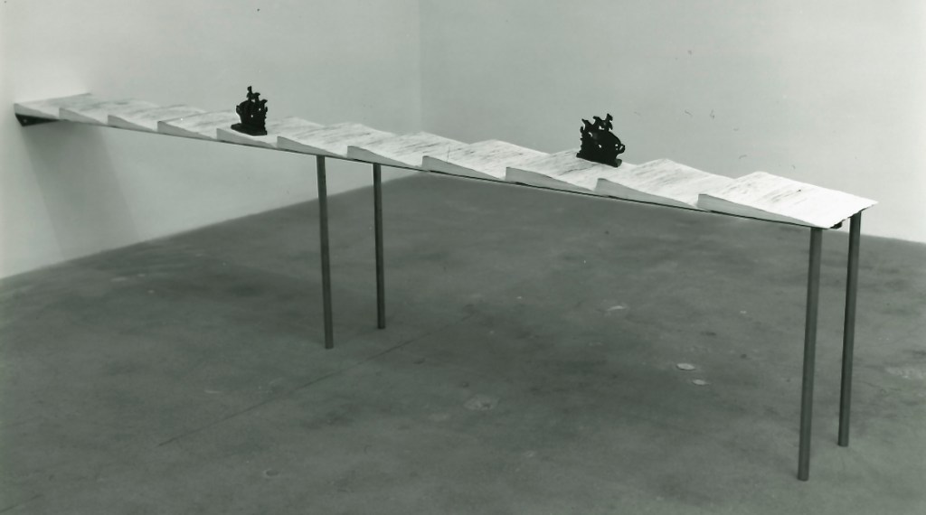 A minimalist art installation featuring a long table made of flat surfaces resembling open books, with black sculptural pieces shaped like ships positioned on top.