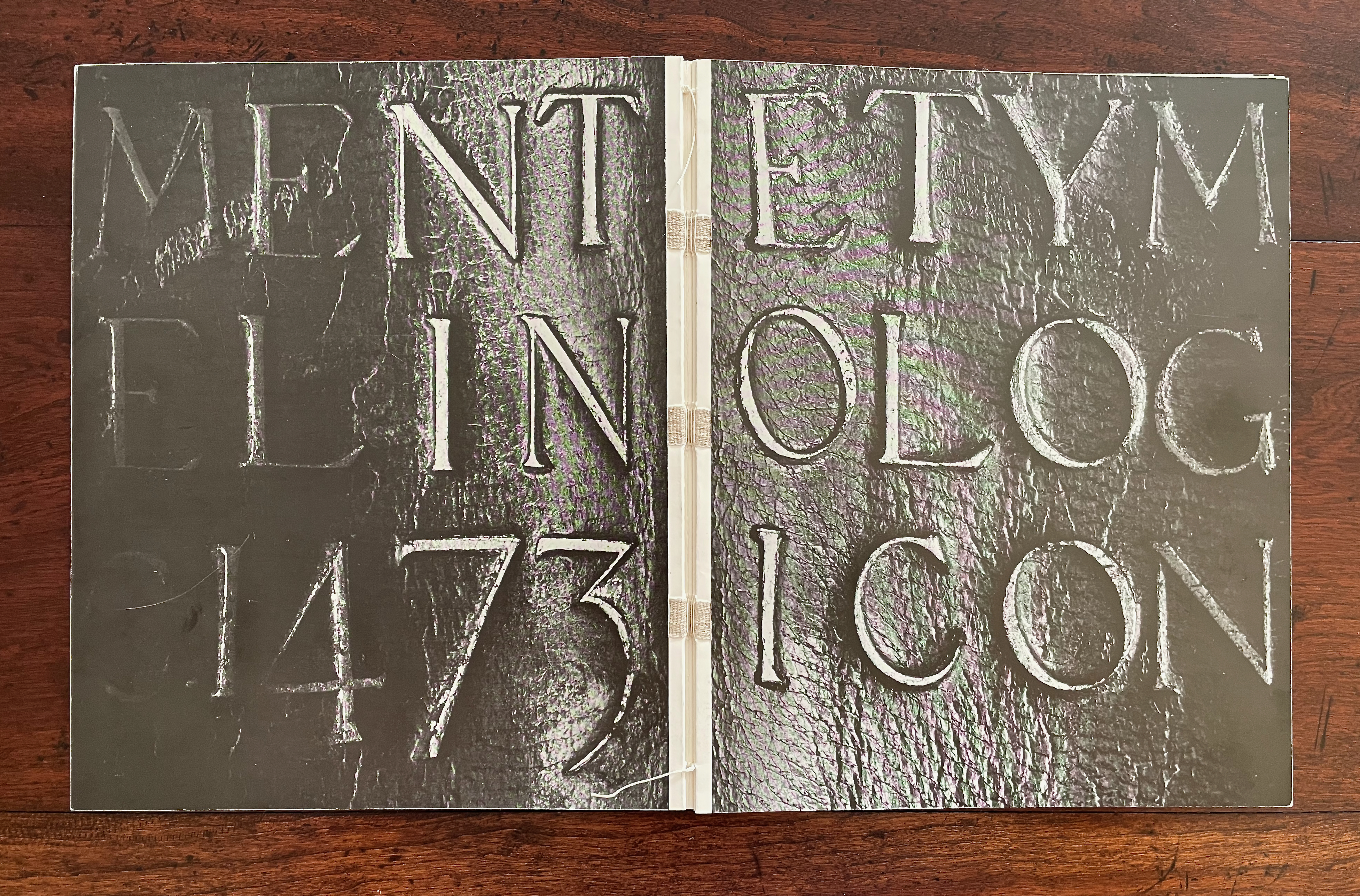 Open book displaying embossed text, including the words 'MENT', 'ETYM', 'EL IN', 'OLOG', and 'ICON', on a dark background.