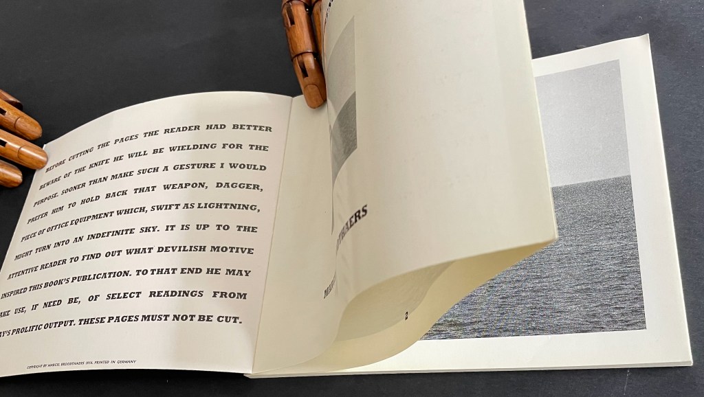 An open book viewed from above, with one page turning and revealing text on the left side and a black and white image of water on the right side. A wooden hand model is holding the book.