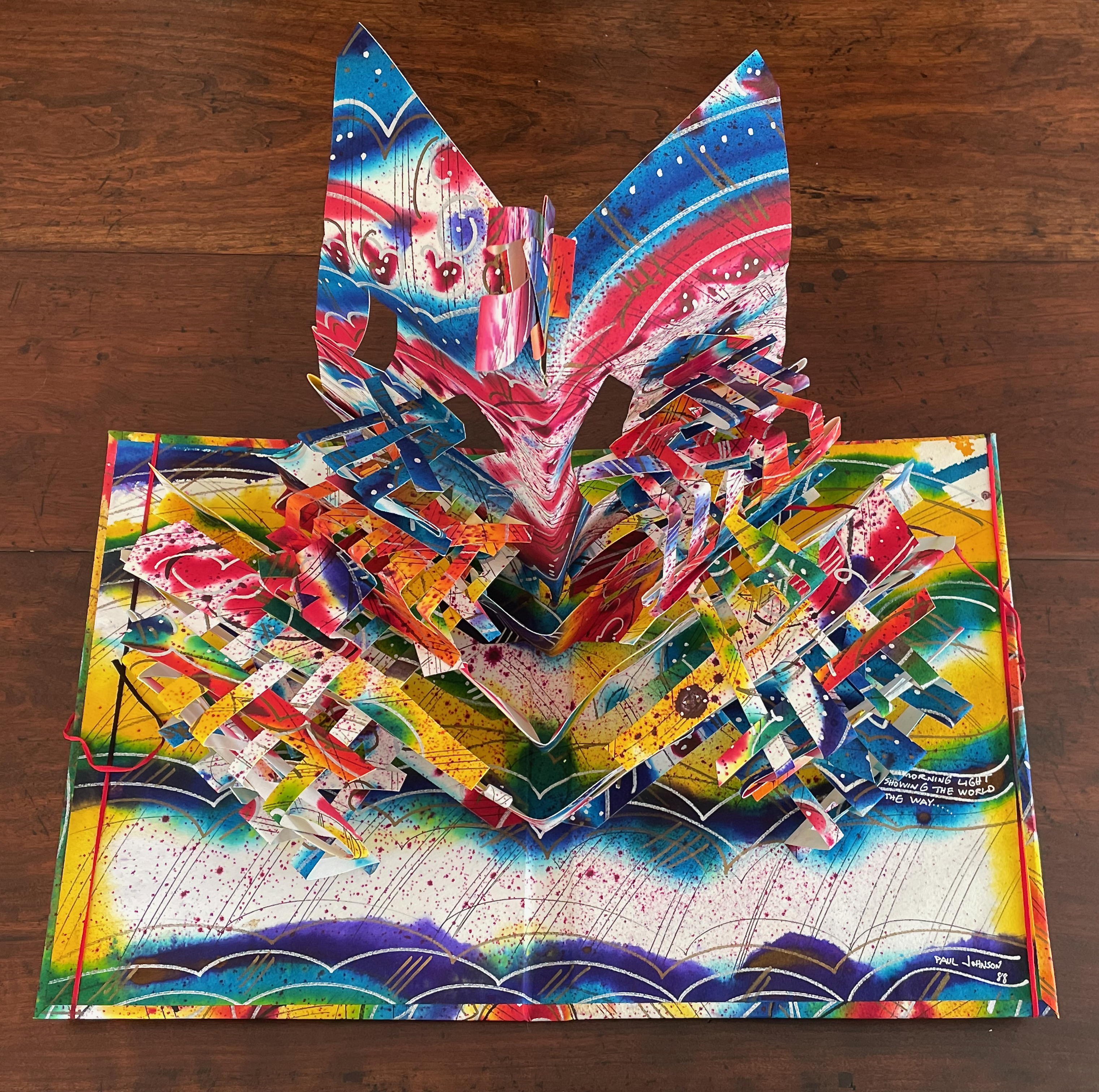 A colorful pop-up book featuring intricate paper designs with vivid patterns and textures. The artwork includes bright hues of red, blue, yellow, and white, creating a visually striking three-dimensional effect.