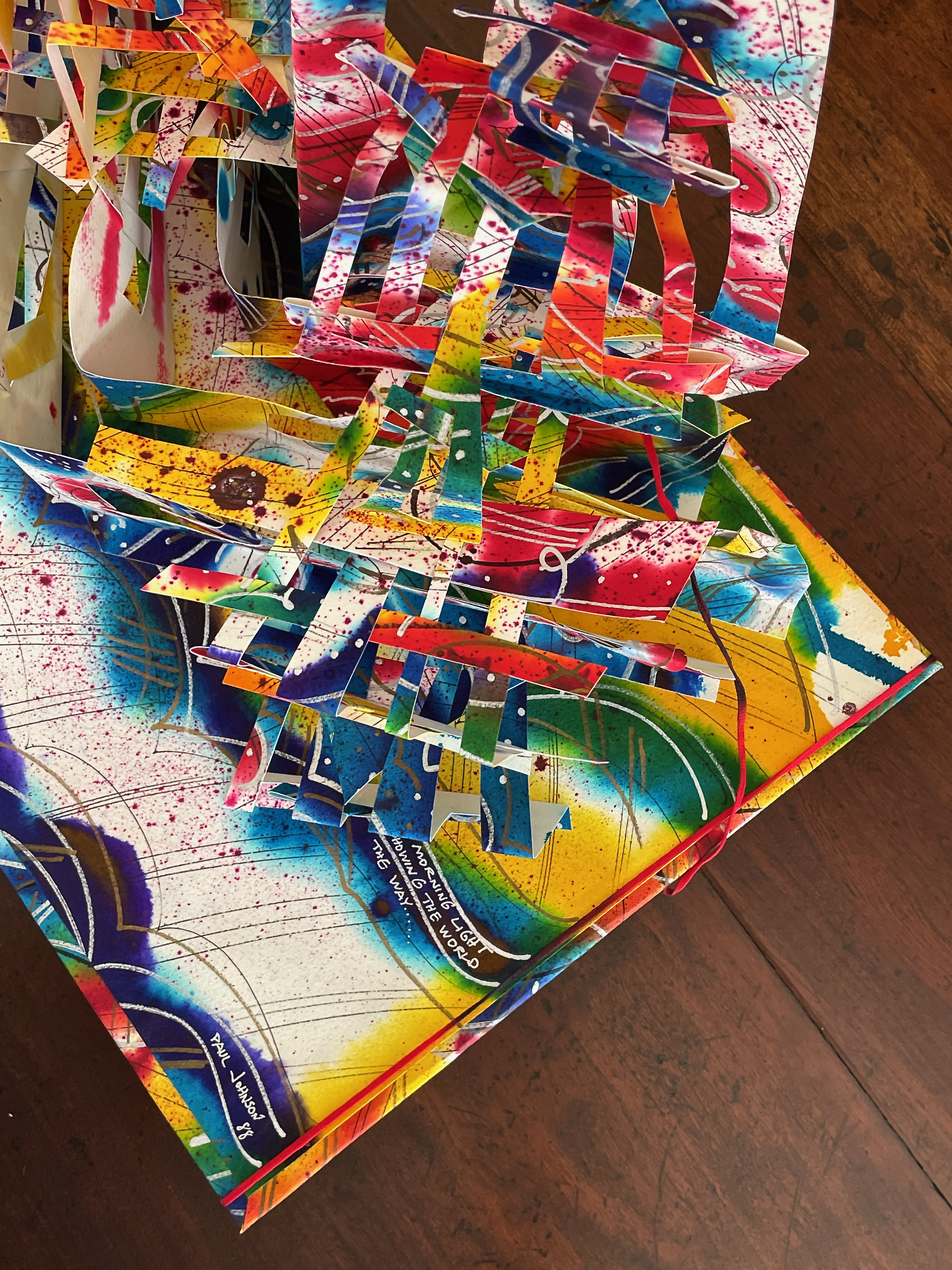 A colorful pop-up art piece featuring vibrant layered paper cutouts in various shapes and patterns, resting on a wooden surface.