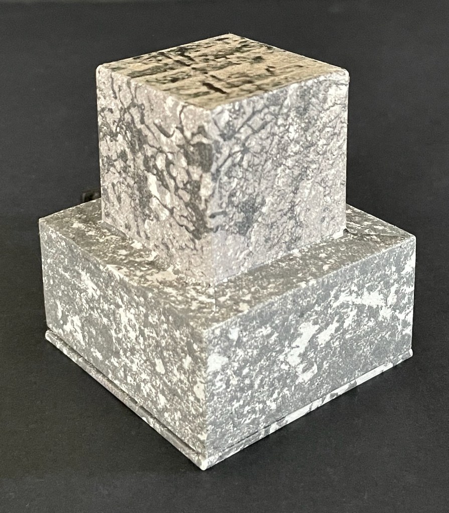 A decorative two-tier sculpture made of textured gray stone, featuring a smaller square block resting on a larger square base.