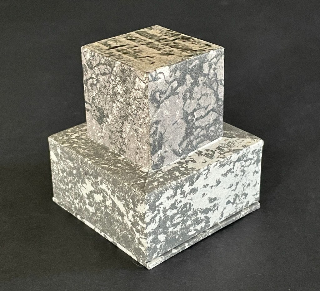 A stacked geometric sculpture with a textured surface, featuring a smaller square top and a larger rectangular base in shades of gray.