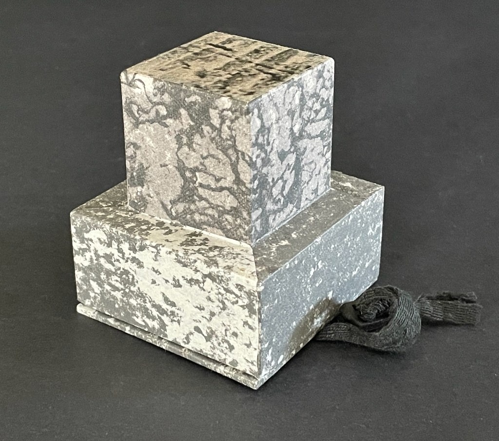A two-layered stone sculpture featuring a square base and a smaller square top, both with a mottled gray and white pattern, resting on a black surface.