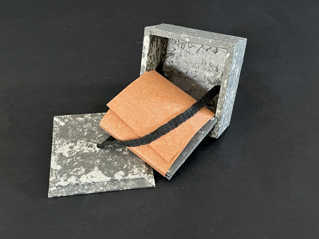 An open decorative box made of textured gray material containing small orange booklets tied with a black string.