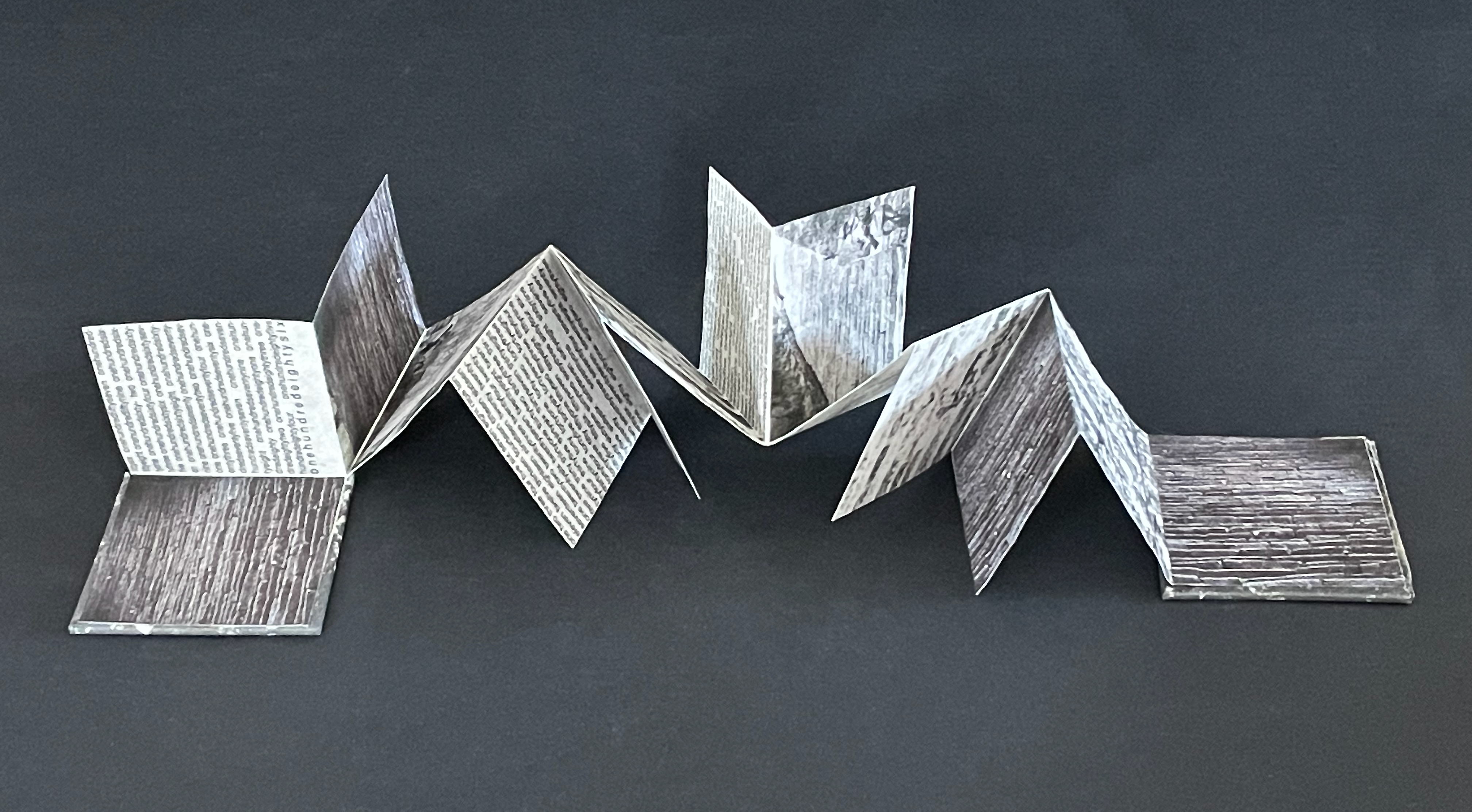An artistic fold-out book made of paper, displaying various textures and printed text, arranged on a dark background.