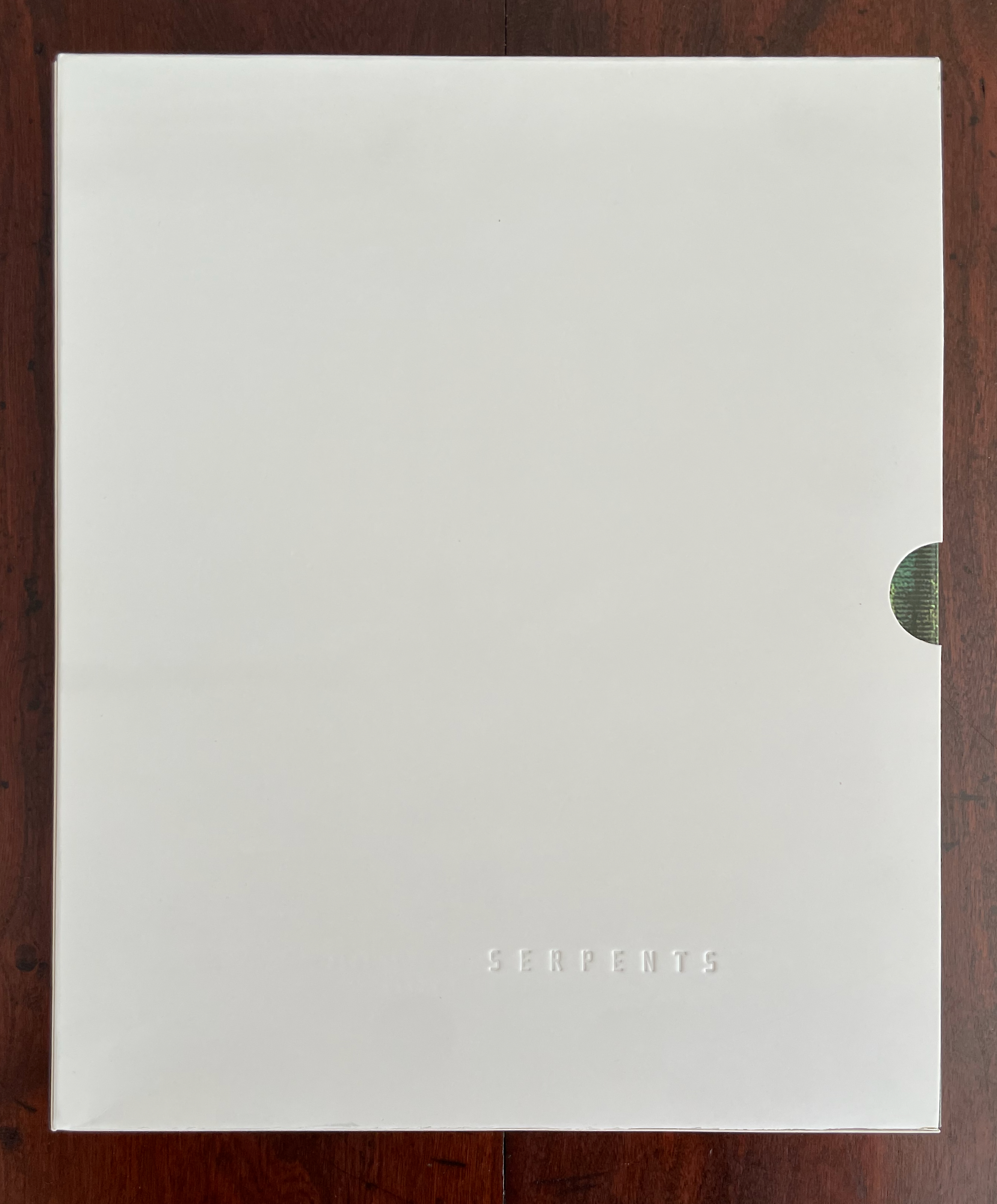 A closed white folder with the word 'SERPENTS' embossed on the front, resting on a wooden surface.