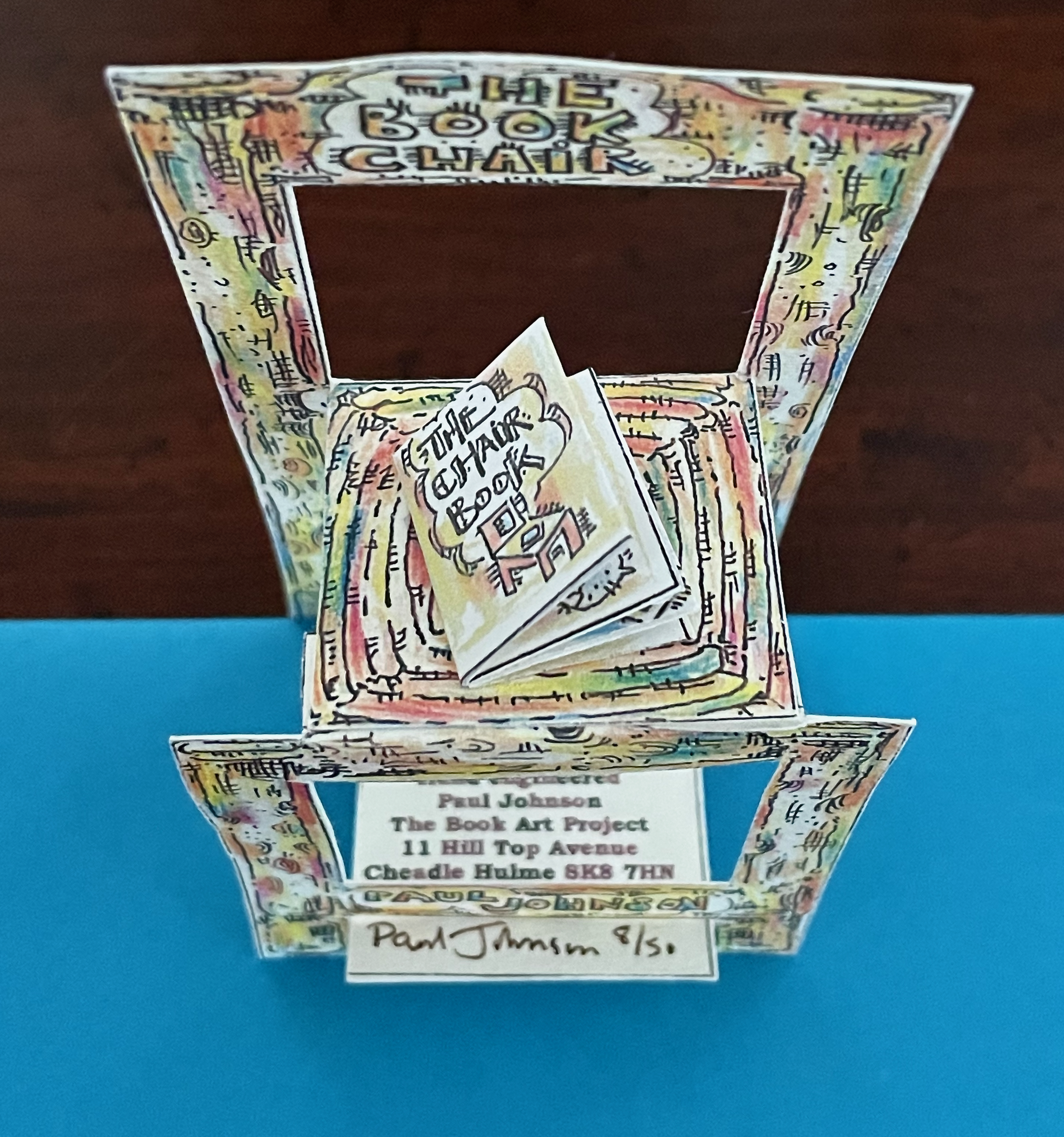 A colorful paper sculpture resembling a chair with a book on its seat, labeled 'The Book Chair'. The sculpture features intricate designs and includes a card with the artist's name, Paul Johnson, and project details.