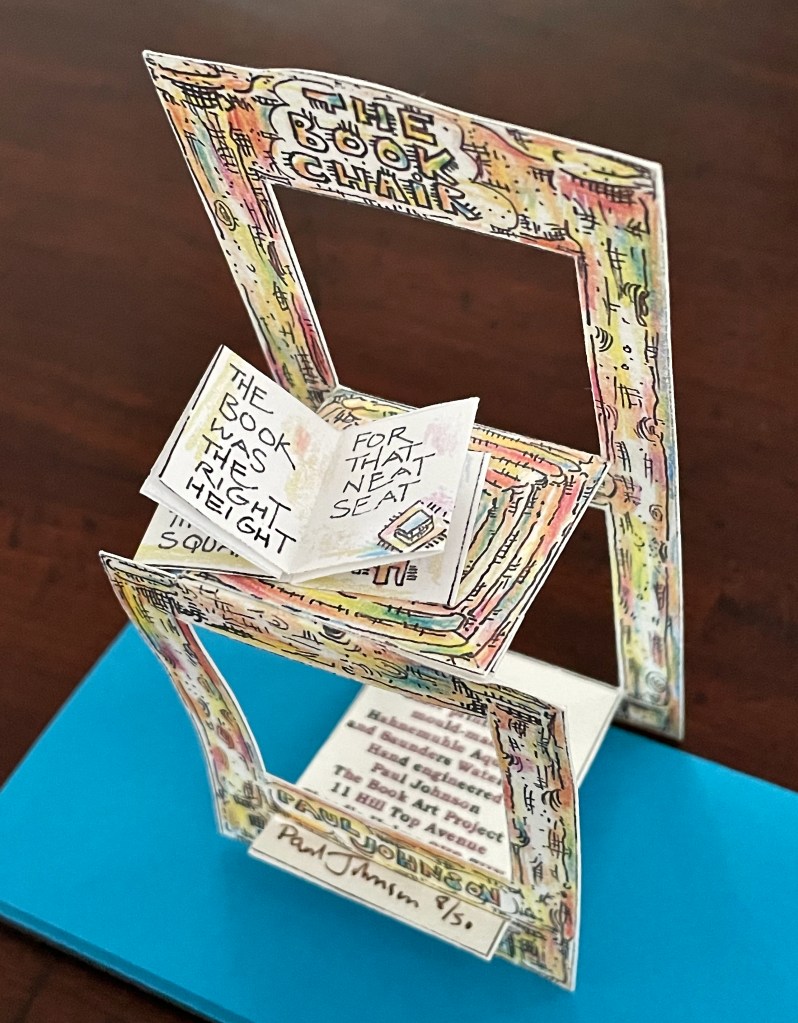 A colorful, paper-crafted model of a chair titled 'The Book Chair' with an open book displayed on it, featuring hand-drawn text and illustrations.