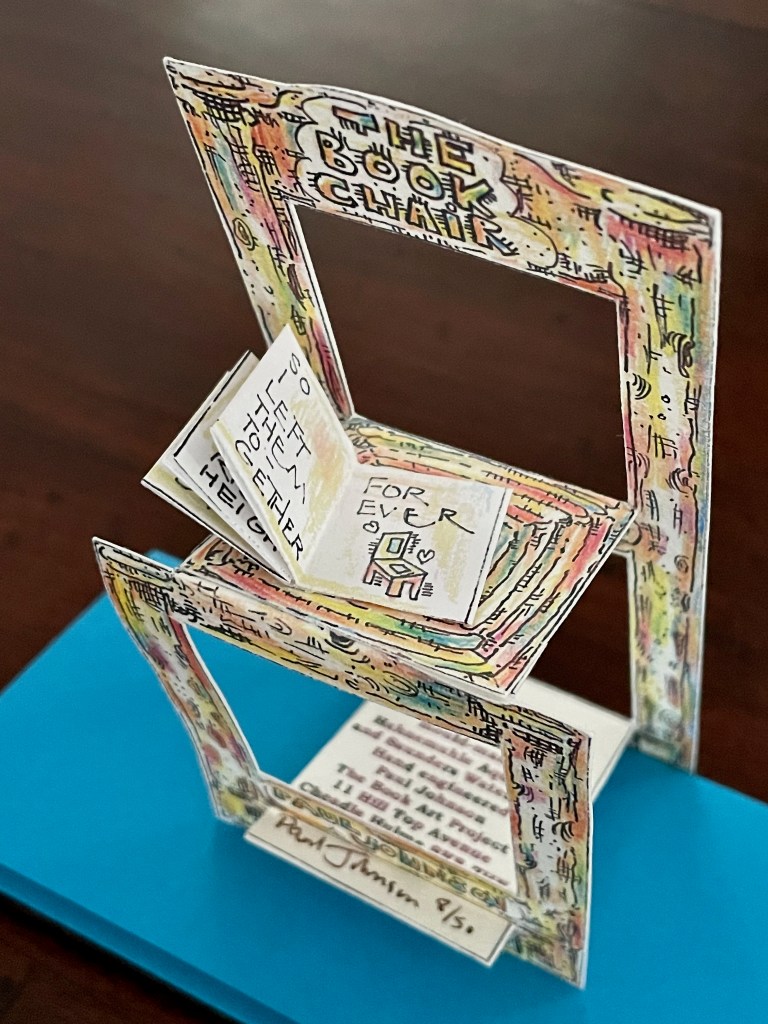 A colorful pop-up art piece titled 'The Book Chair,' featuring intricately illustrated layers of a chair and books, with handwritten text elements and a blue base.