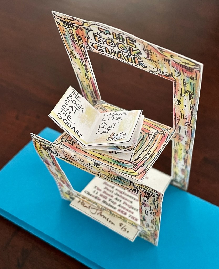 A colorful pop-up card featuring a chair and an open book with playful text, set against a blue background.